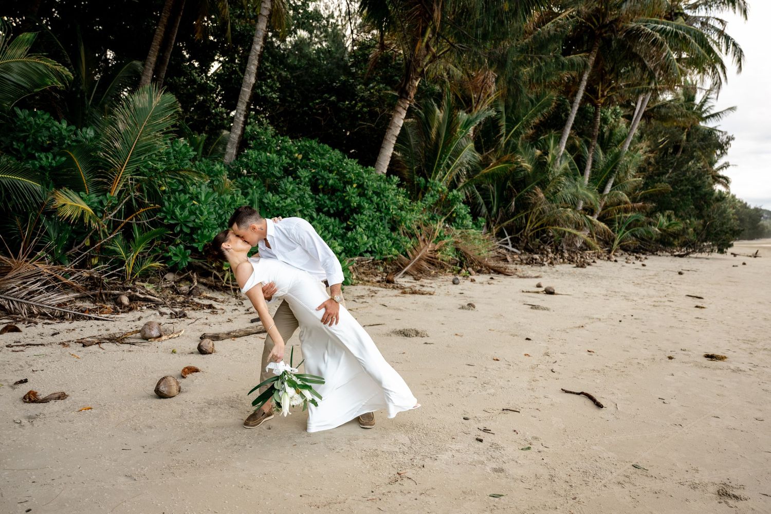Groom dips bride for a kiss on sandy tropical beach with palm trees in background at Four Mile Beach, Port Douglas.