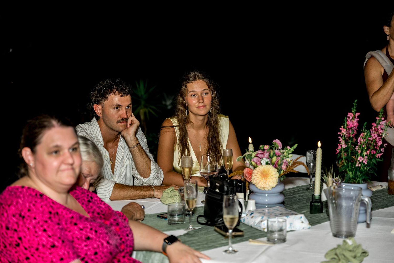 Couple and guests seated at a candlelit dinner table with floral centerpieces at a night event.