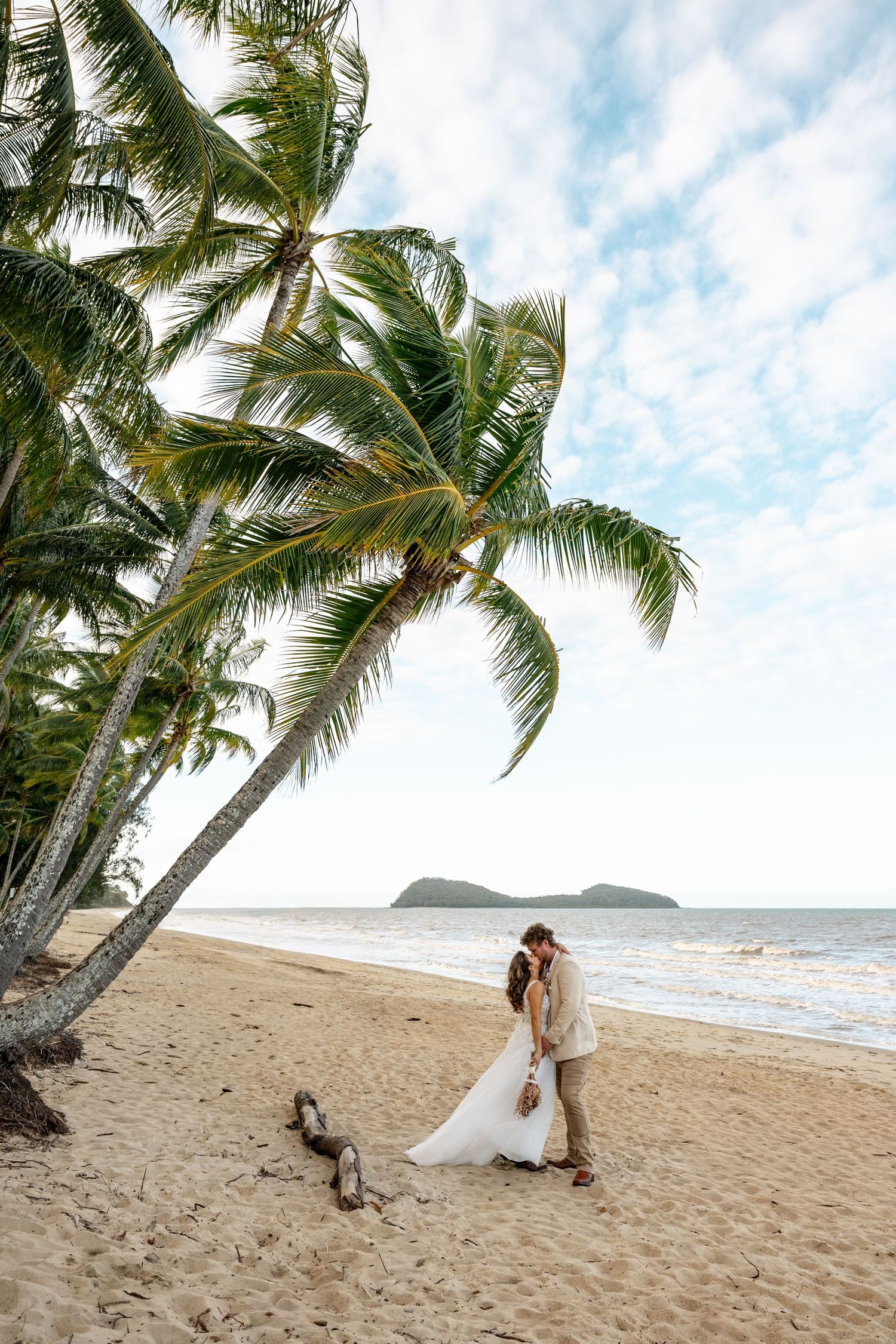 Bride and groom share a romantic kiss under a leaning palm tree on a tropical sandy beach.