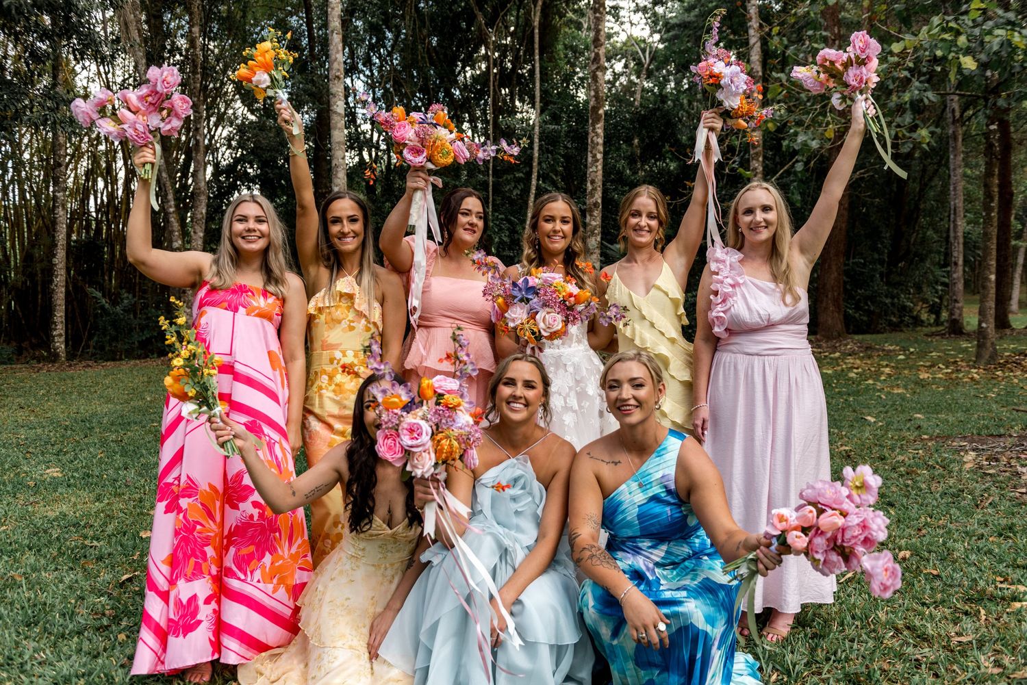 Bridesmaids in colorful dresses raising bouquets with bride at outdoor forest wedding celebration.