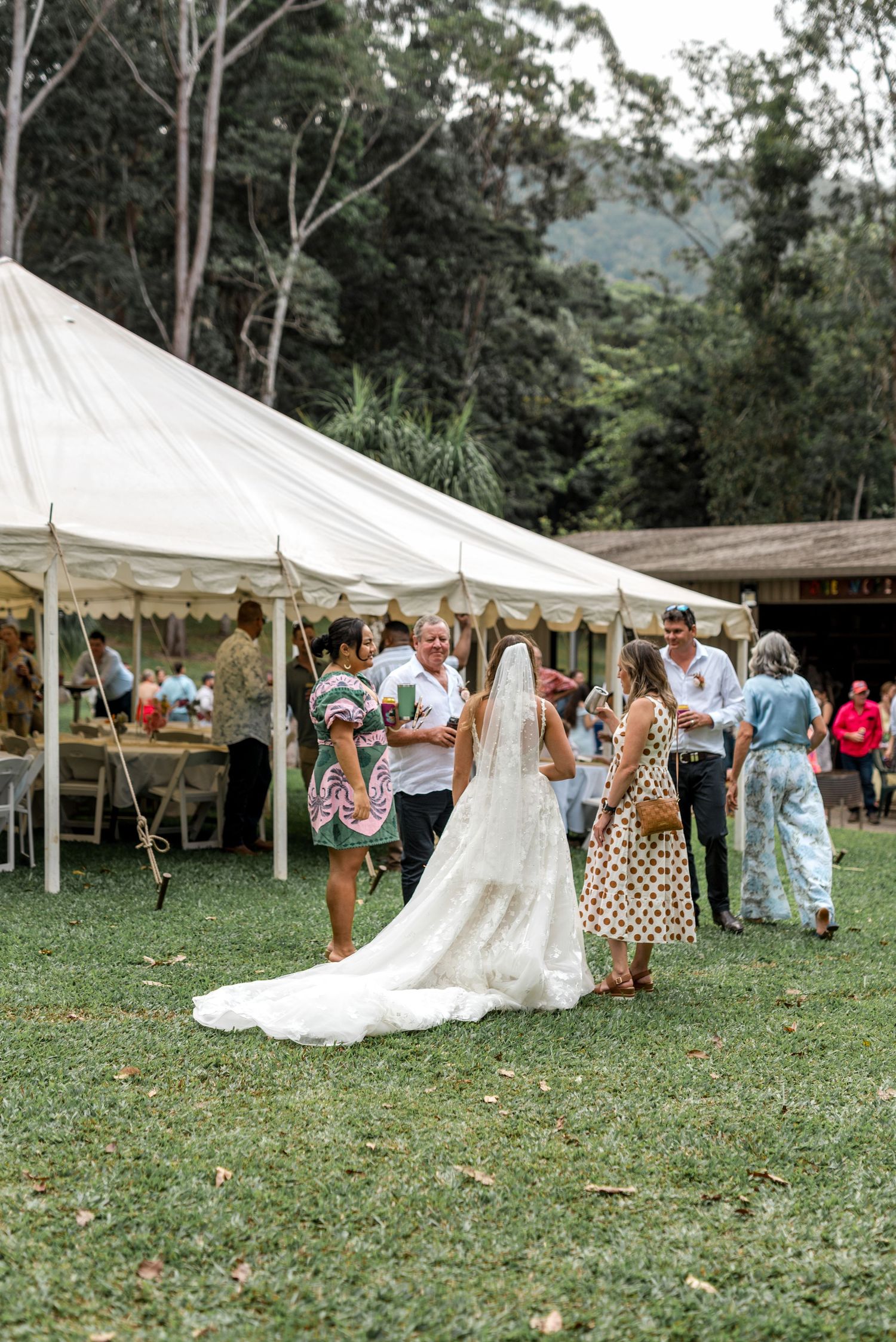 Bride in white gown with long veil mingles with guests near a large white marquee tent outdoors.