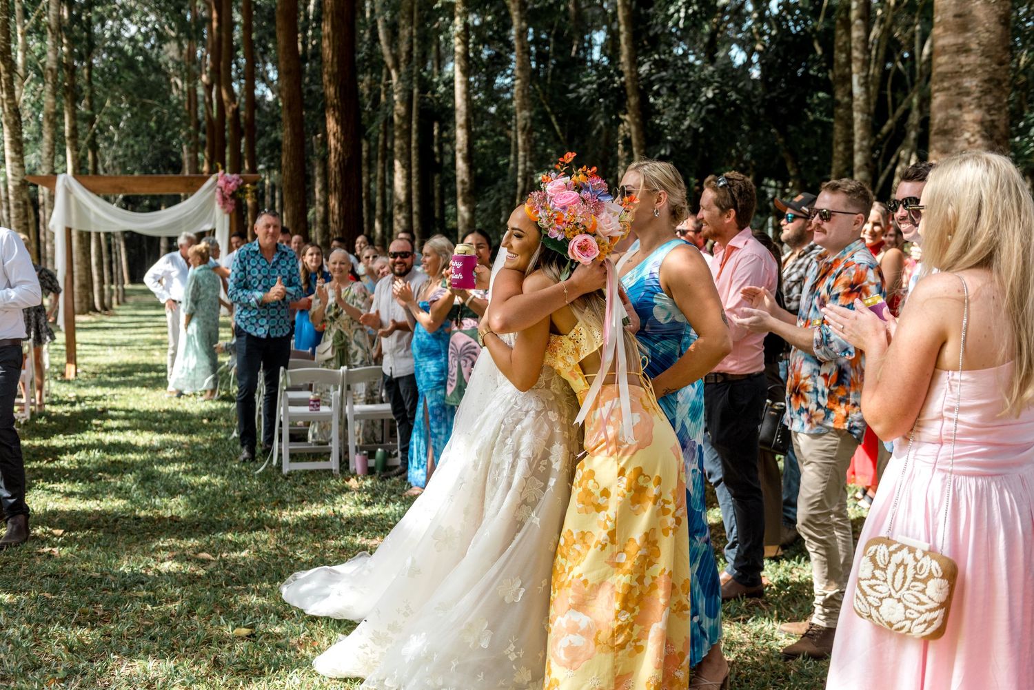 Bride embraces a guest in a floral dress while holding a bright bouquet during outdoor woodland wedding celebration.