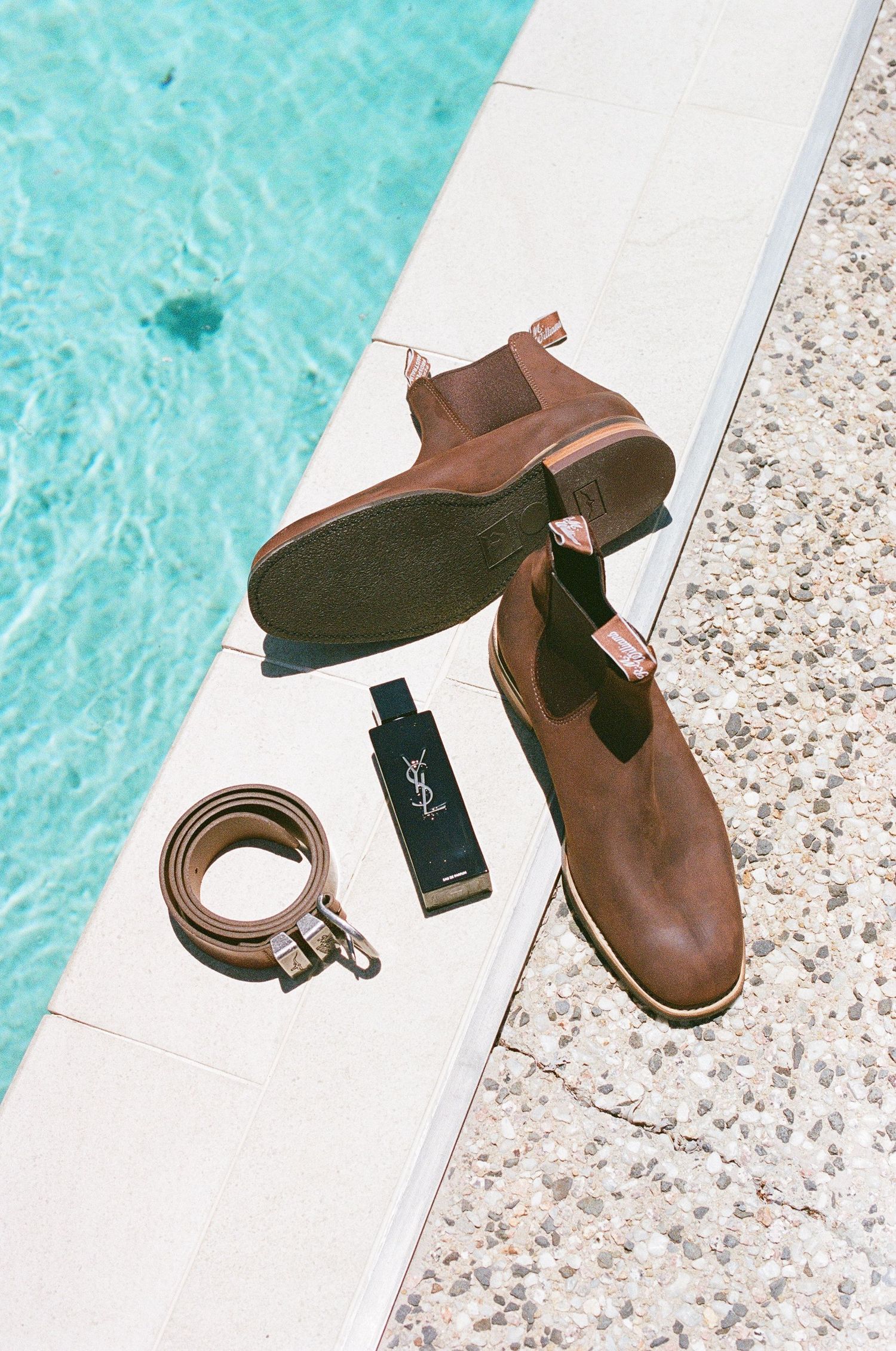 Brown leather Chelsea boots, YSL cologne, and belt arranged by turquoise pool for groom.