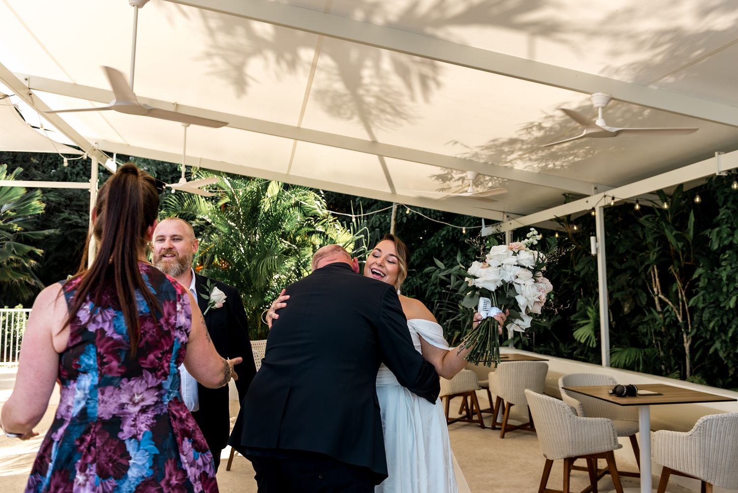 Bride in white gown hugs a guest while holding a white bouquet under a canopy at her wedding.