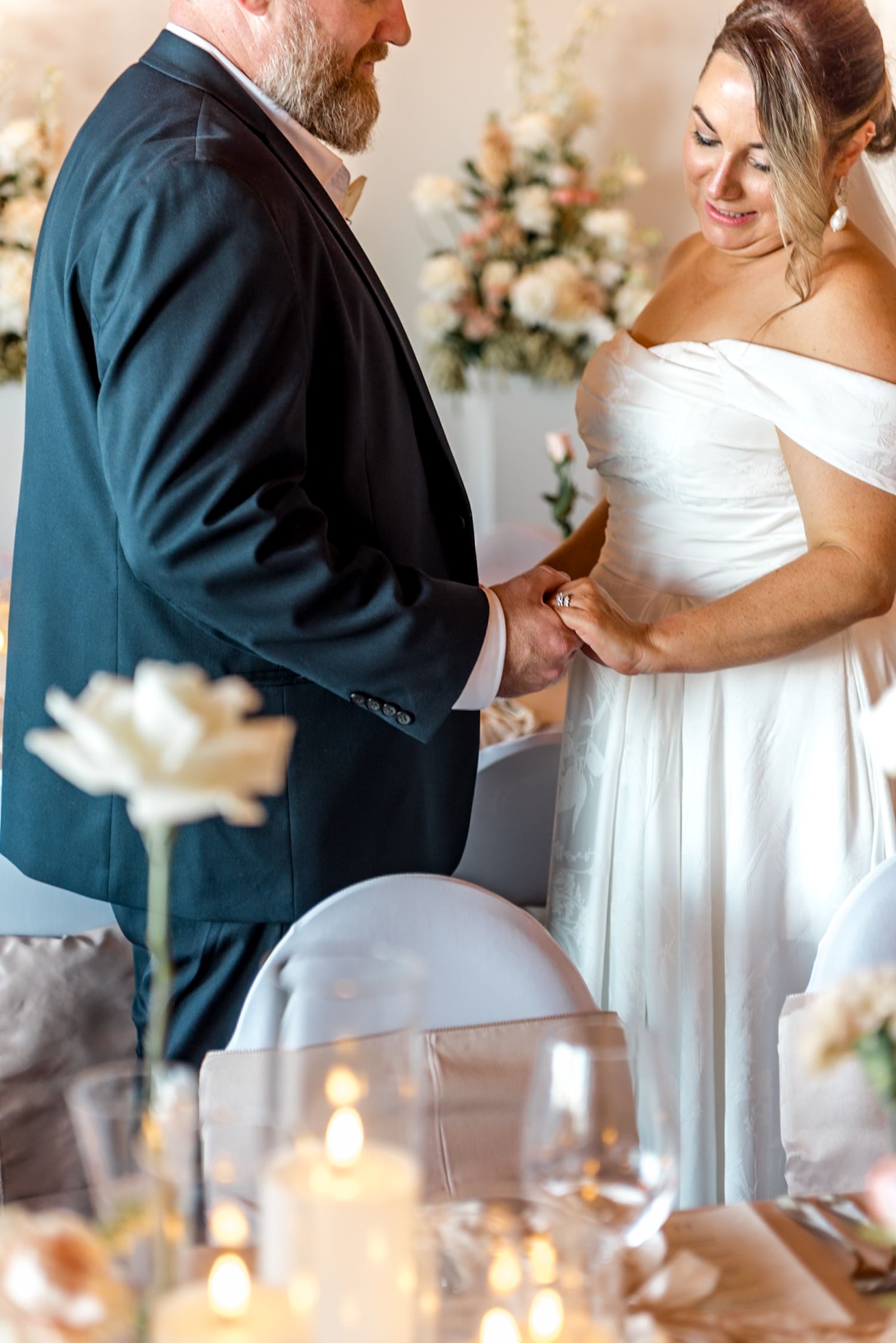 Bride smiles warmly while holding groom's hands at their elegant wedding reception with candle decor.