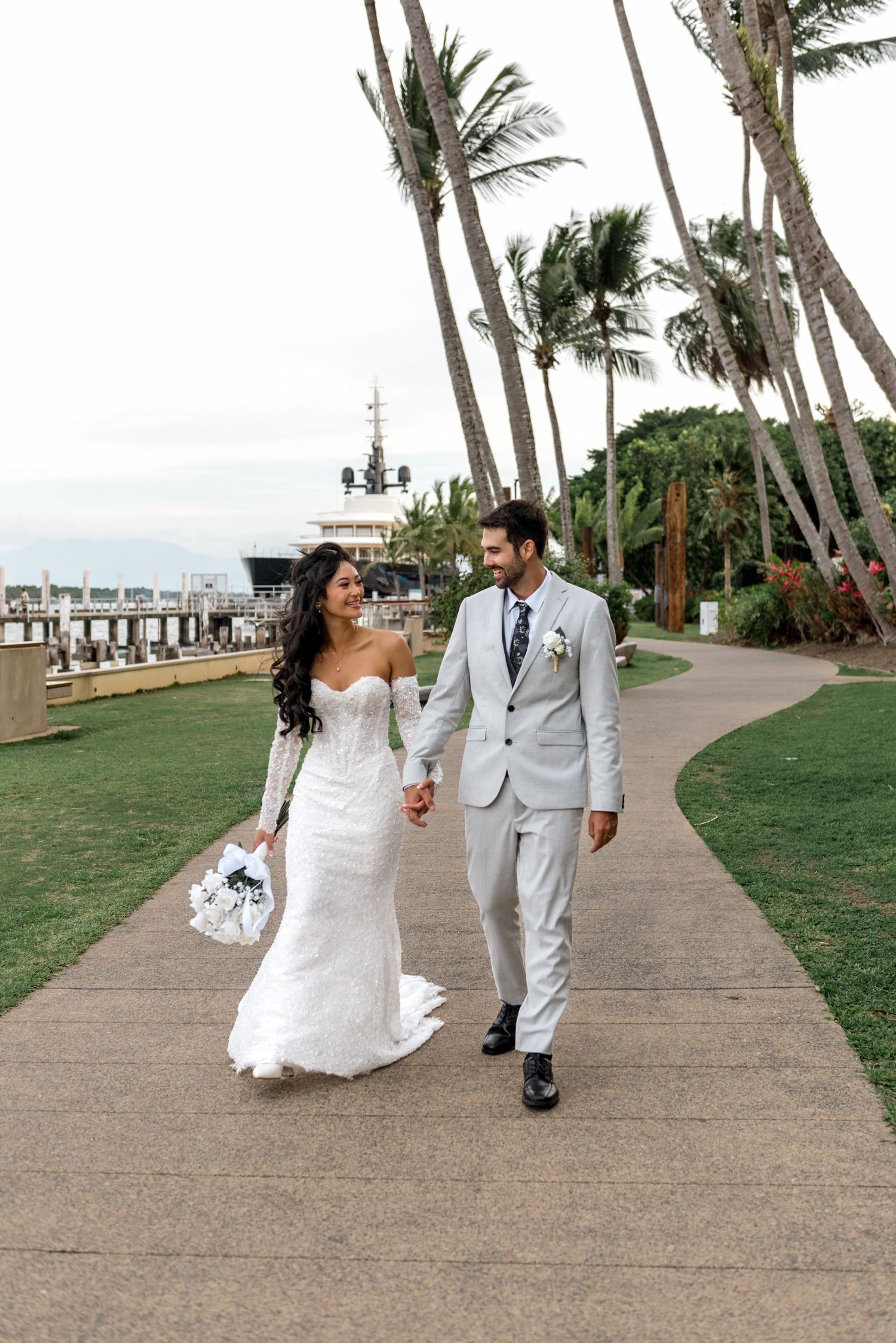 Happy bride and groom walk hand-in-hand along palm-lined waterfront path on their wedding day.