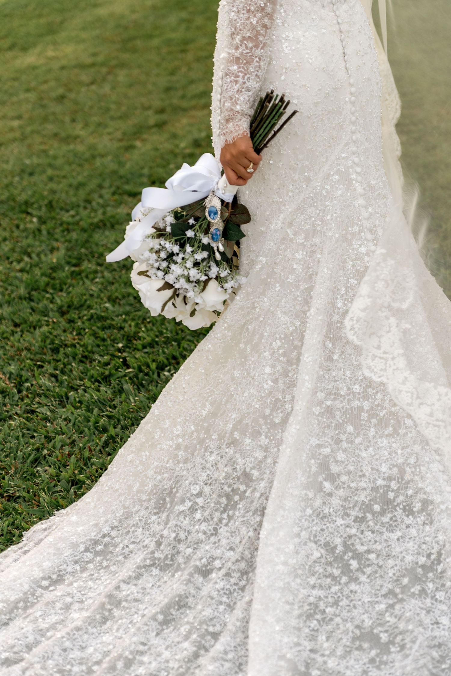 Close-up of bride holding white wedding bouquet with ribbon detail against lace gown train on green grass.