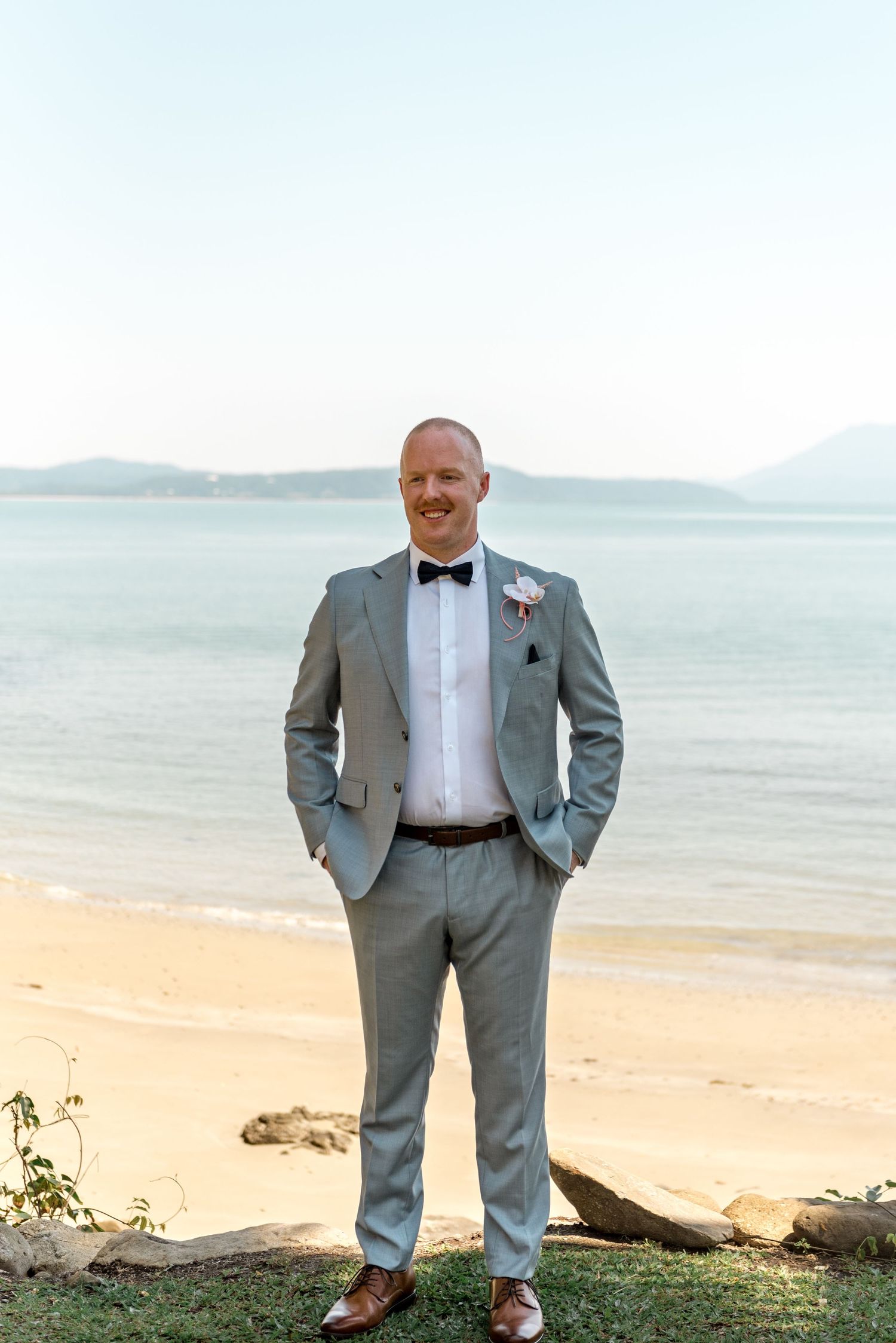 Groom in light gray suit and black bow tie stands smiling on a tropical beach with calm ocean views.