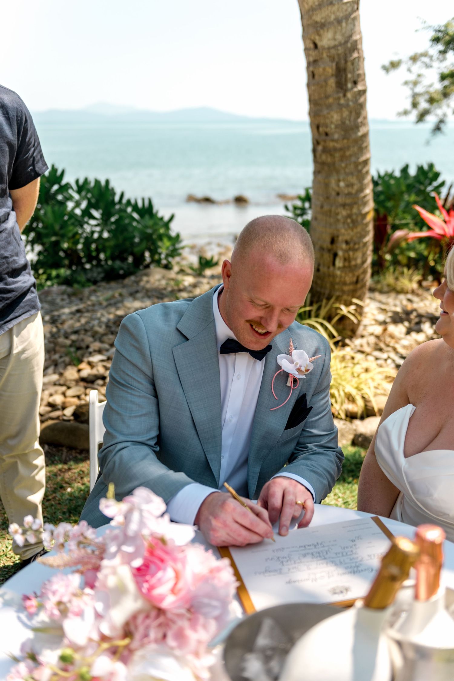 Groom in grey suit signing wedding certificate at outdoor table with ocean views and pink floral arrangements.