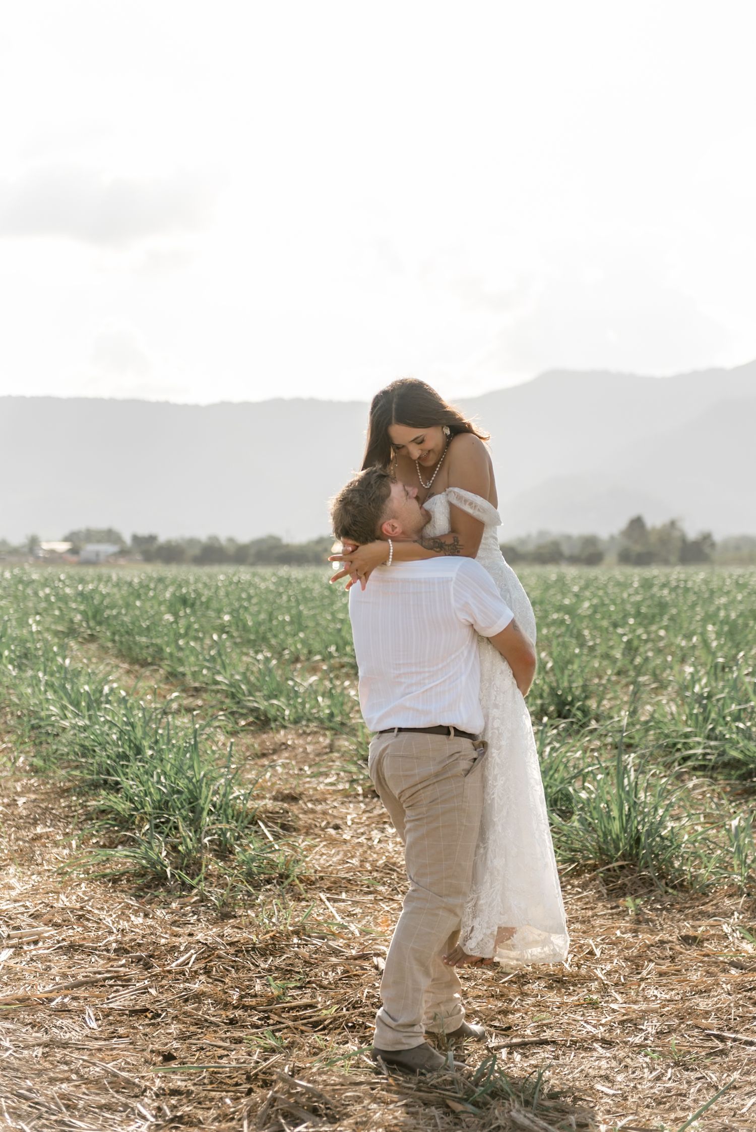 Groom lifting smiling bride in elegant white dress in a wide open green field at their wedding.
