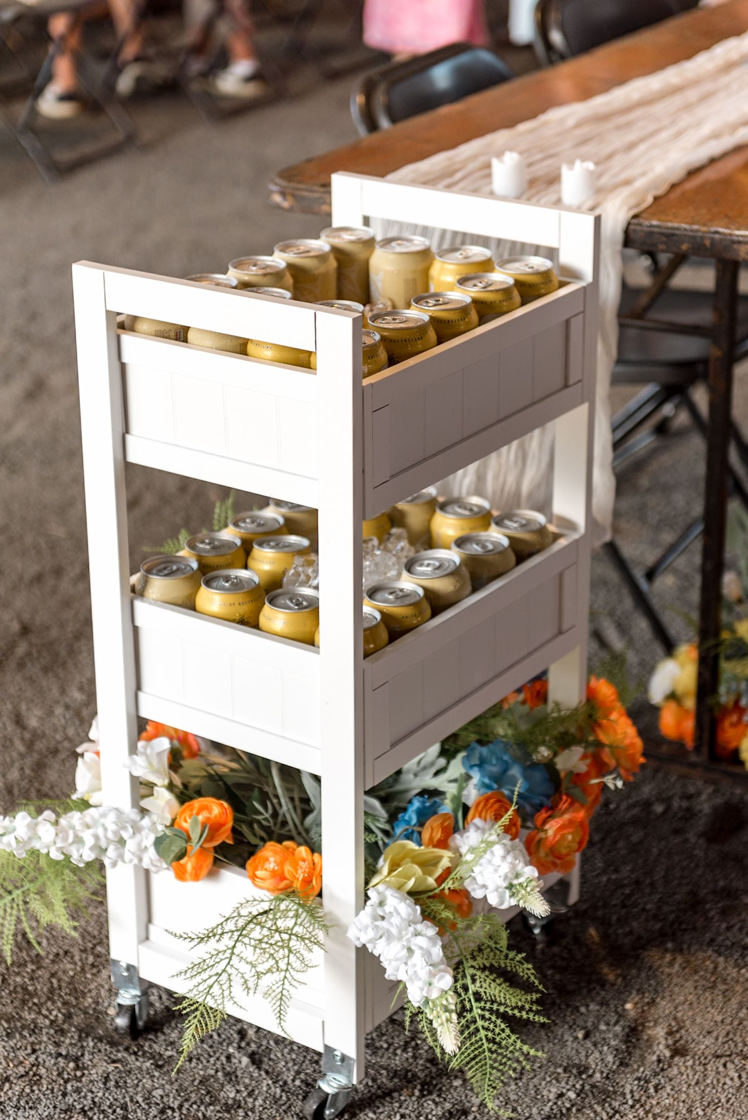 White rolling cart filled with canned beverages and decorated with colorful flowers.