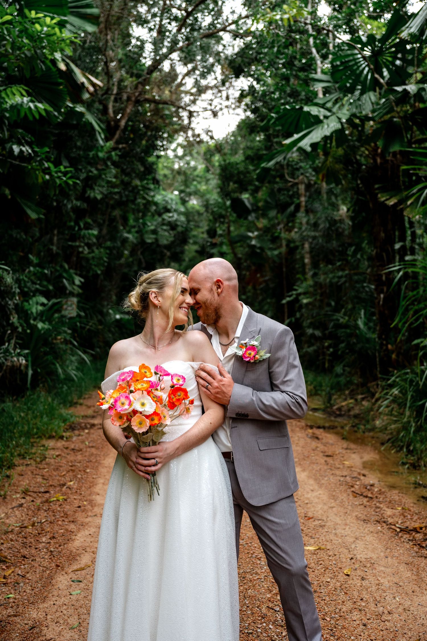 Groom tenderly kisses bride on a jungle dirt path as she holds a bright orange and pink bouquet.