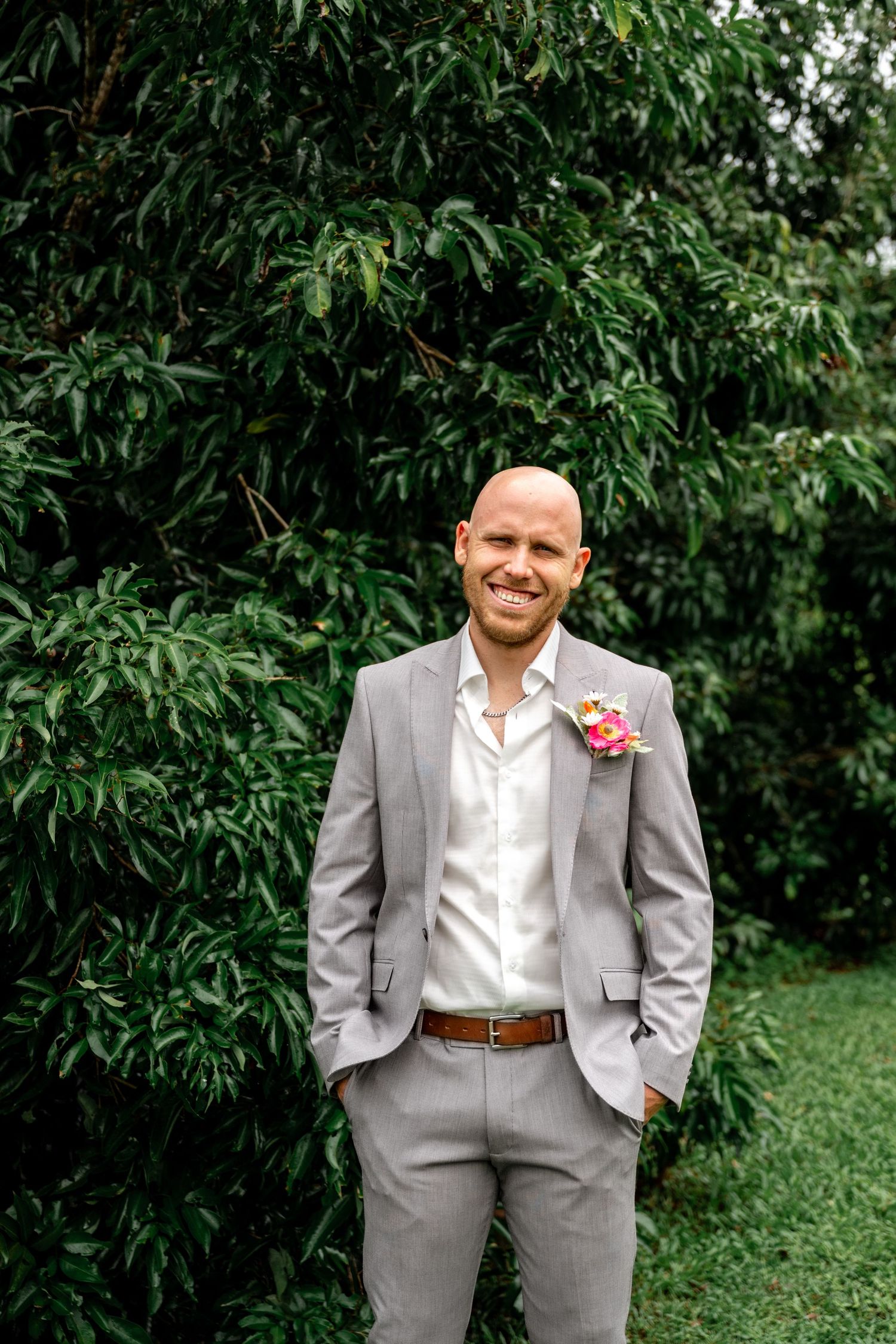Happy bald groom in grey suit with colorful boutonniere smiling outdoors against lush green trees.