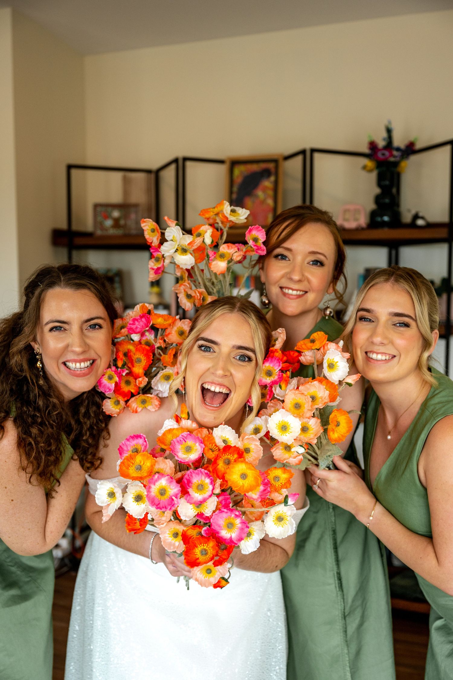 Bride and three bridesmaids in sage green dresses laugh while holding colorful orange and pink flower bouquets indoors.