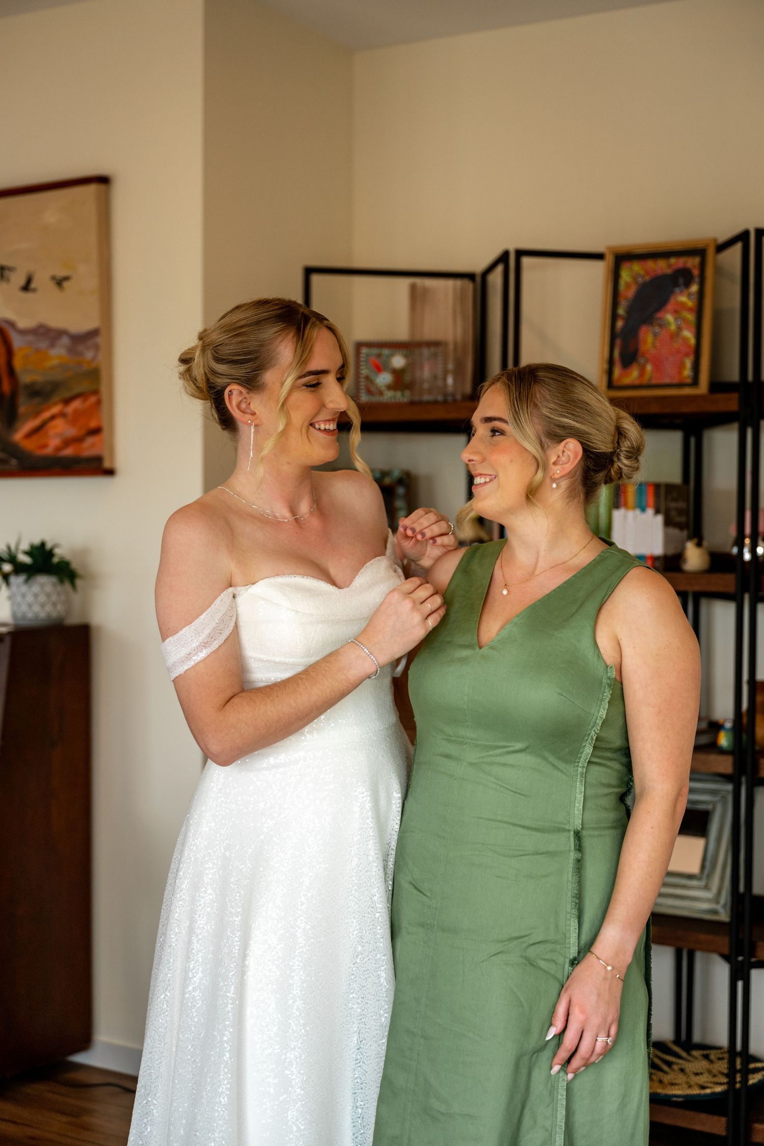 Bride in white gown and bridesmaid in sage green dress share a joyful moment together before wedding.