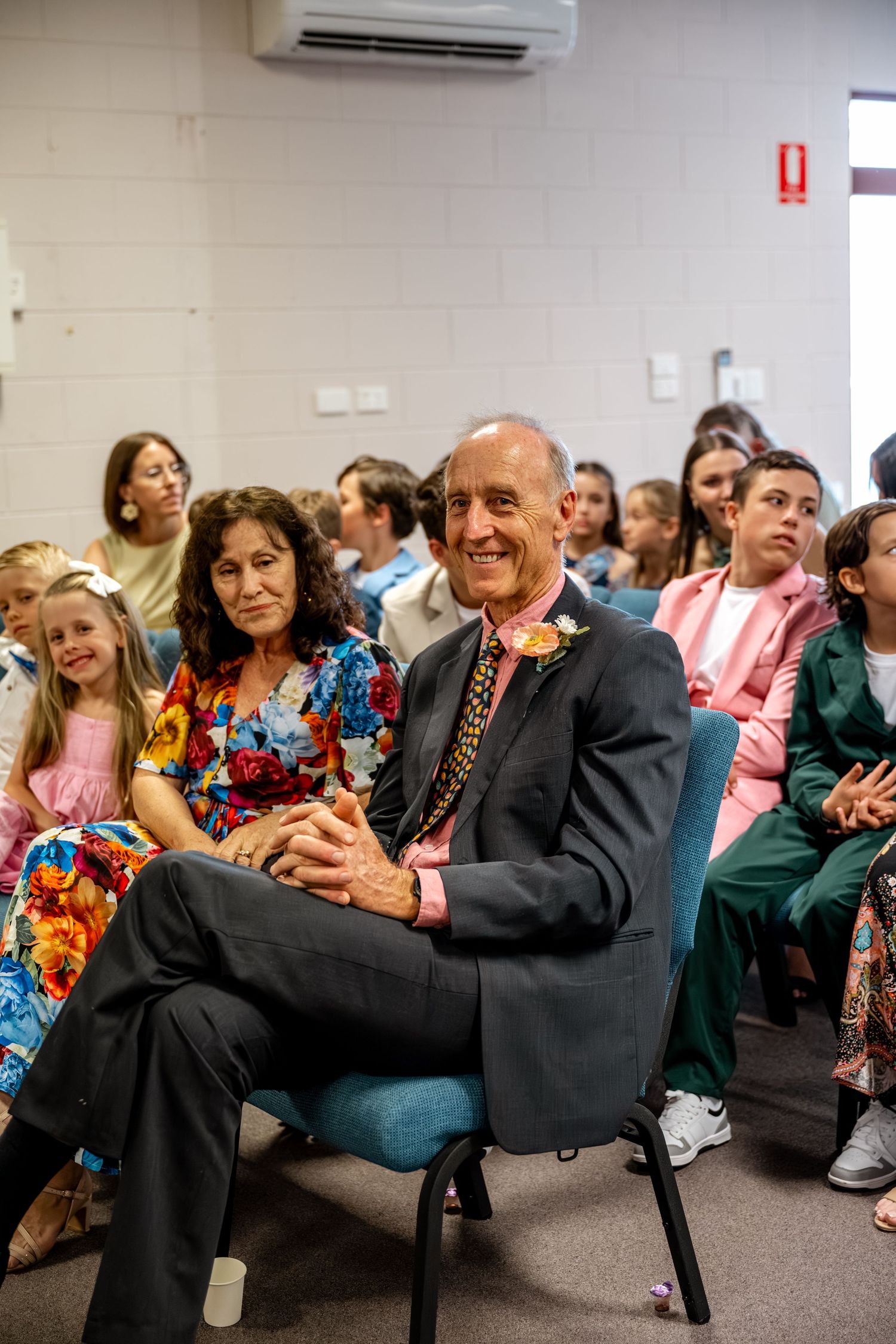 Elegantly dressed wedding guests smile and watch ceremony from teal chairs in indoor venue.