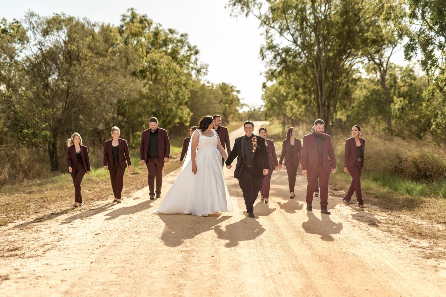 Bride and groom lead wedding party in matching burgundy suits down a rustic rural dirt road.