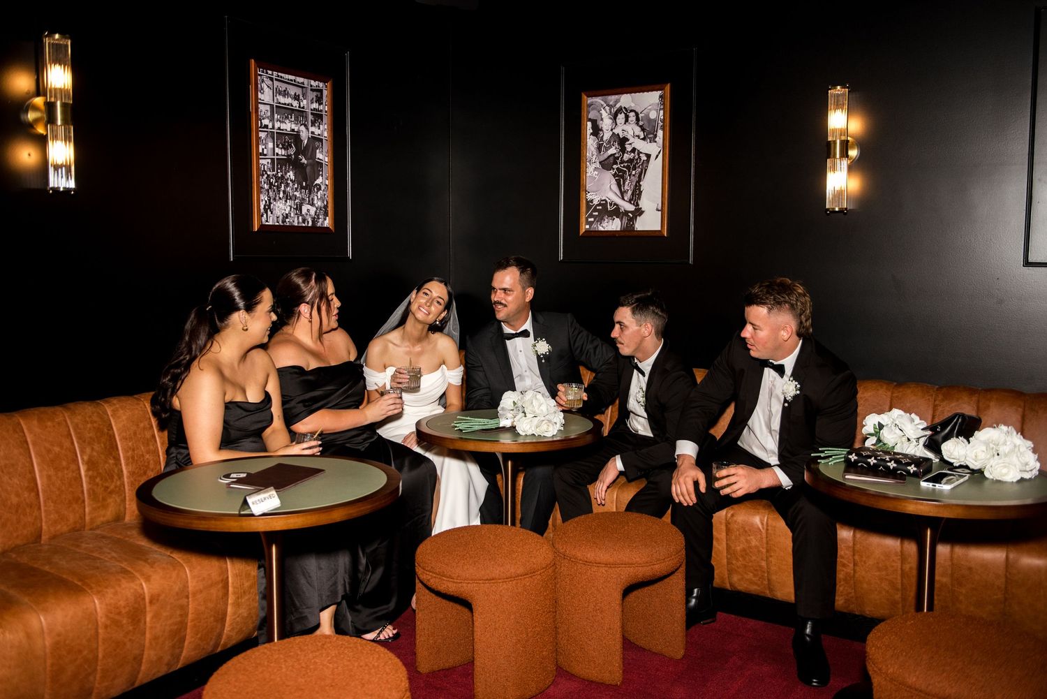 Wedding party relaxes together in a luxurious dark lounge with leather ottomans and gold lighting.