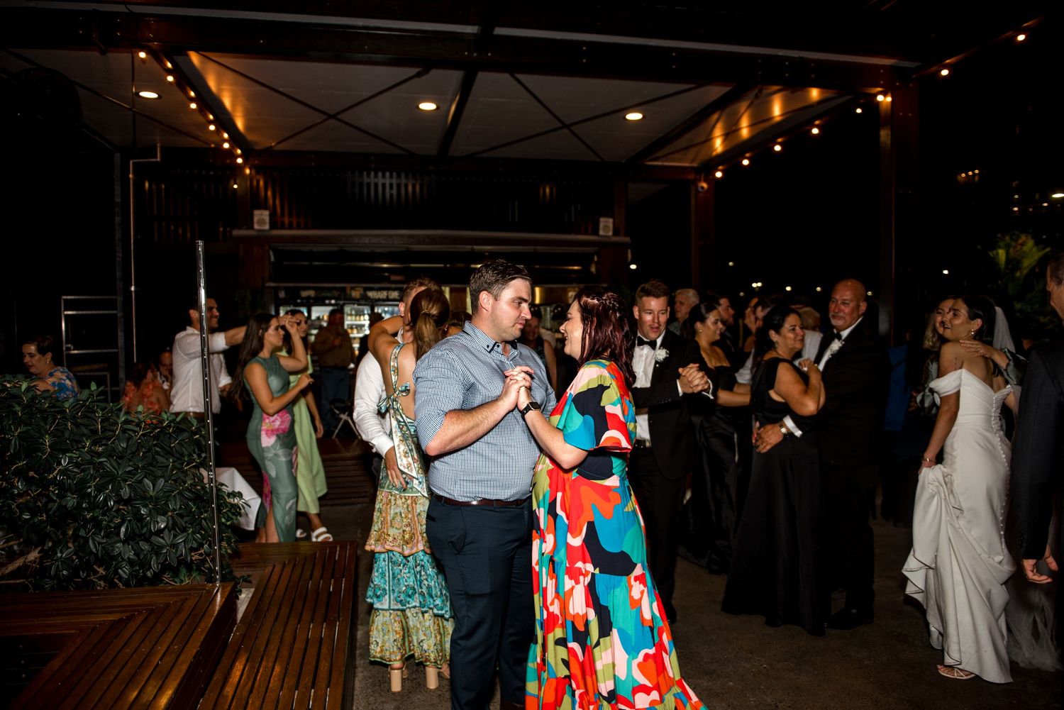 Wedding guests dance joyfully at an outdoor evening reception, including a woman in a colorful floral dress.