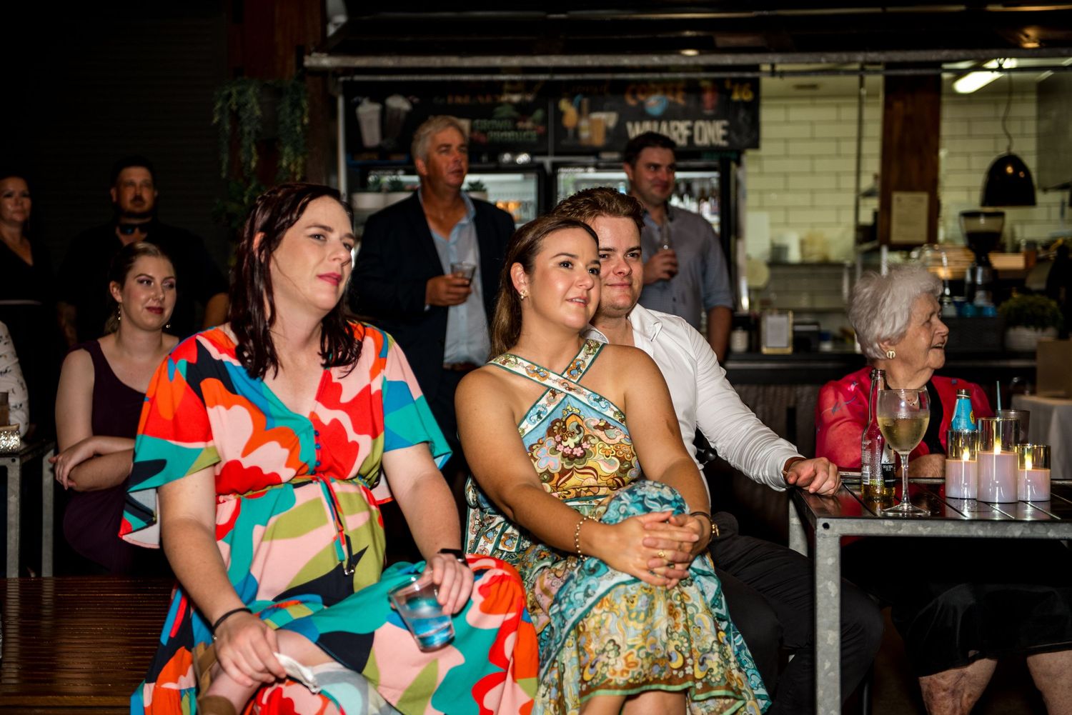 Wedding guests in colorful formal attire smile and watch speeches during an indoor evening reception celebration.
