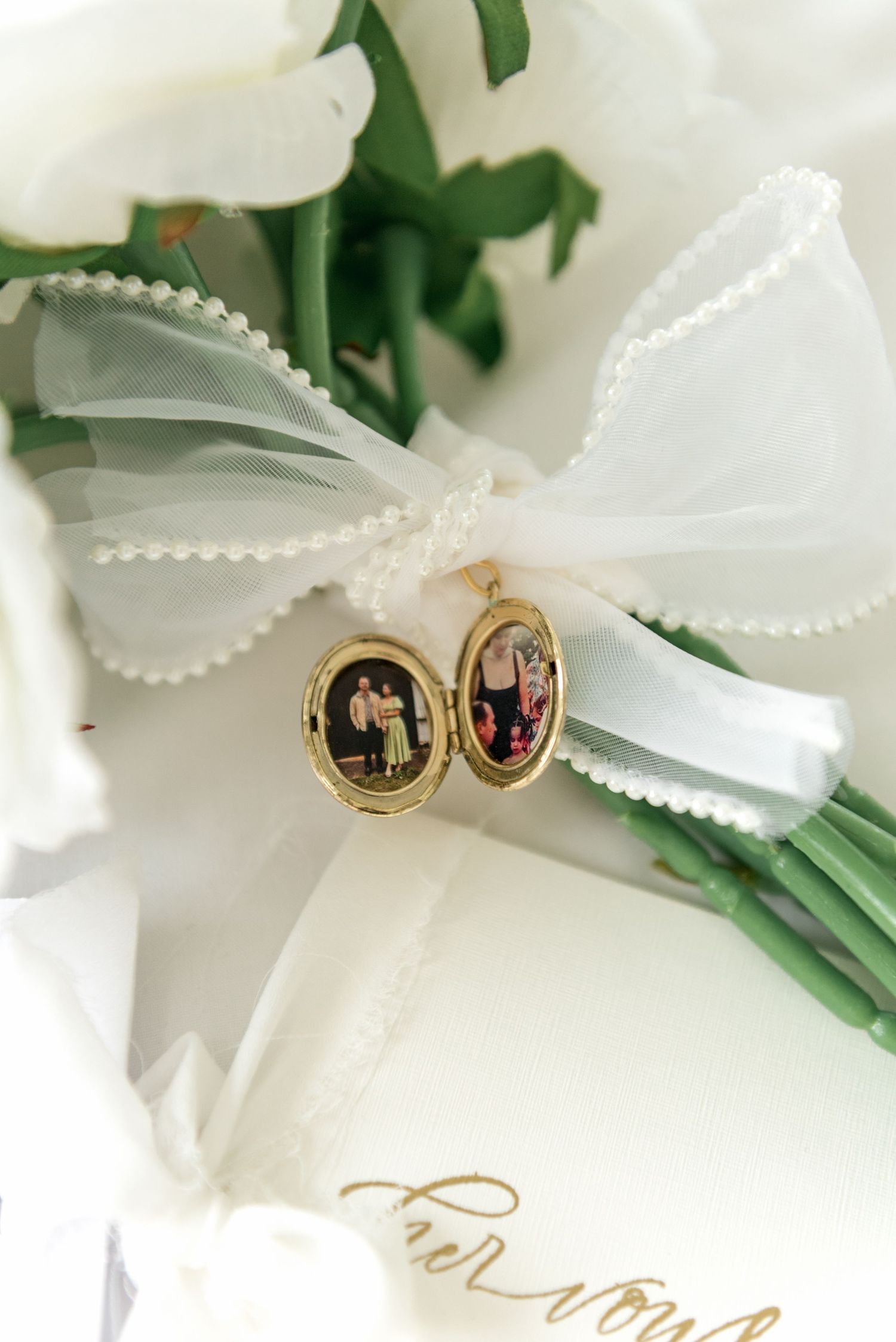Gold locket charm with family photos attached to white ribbon bow on bridal bouquet of white roses.