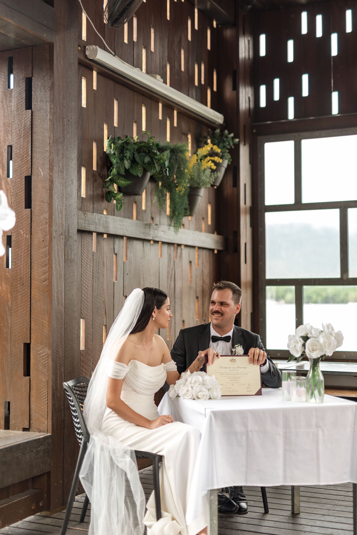 Bride and groom in tuxedo sitting together at decorated table during wedding ceremony indoors.