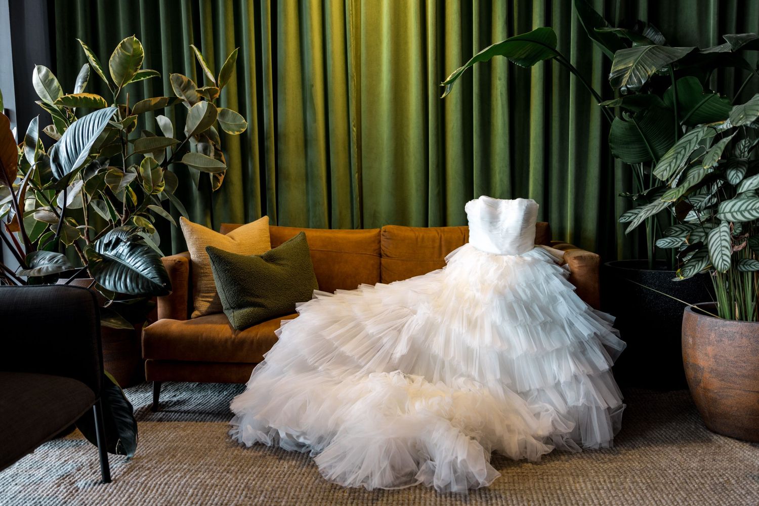 Elegant layered white tulle wedding gown displayed on brown velvet sofa in green styled room.