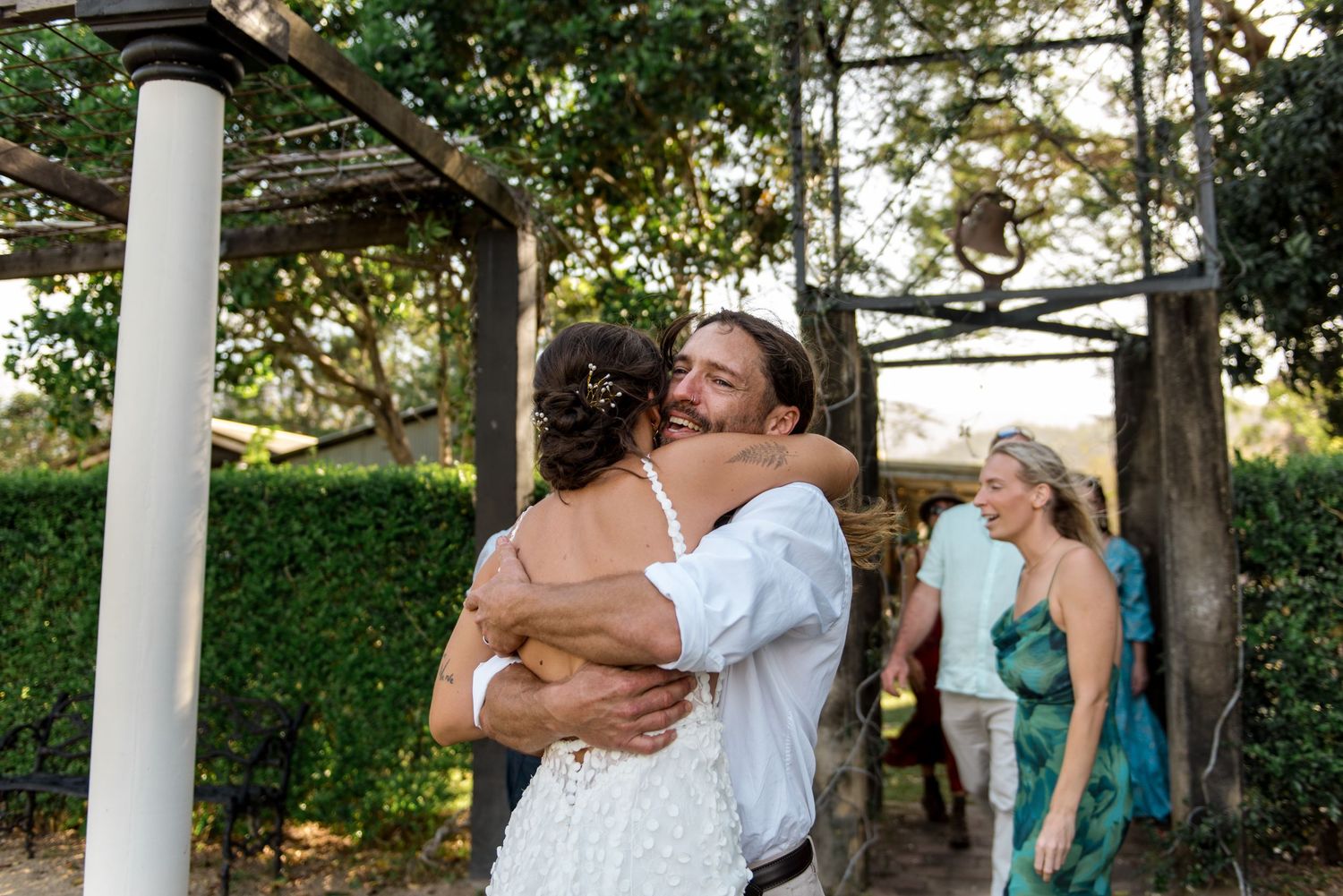 Groom smiling warmly hugs wedding guest under pergola surrounded by lush green trees.