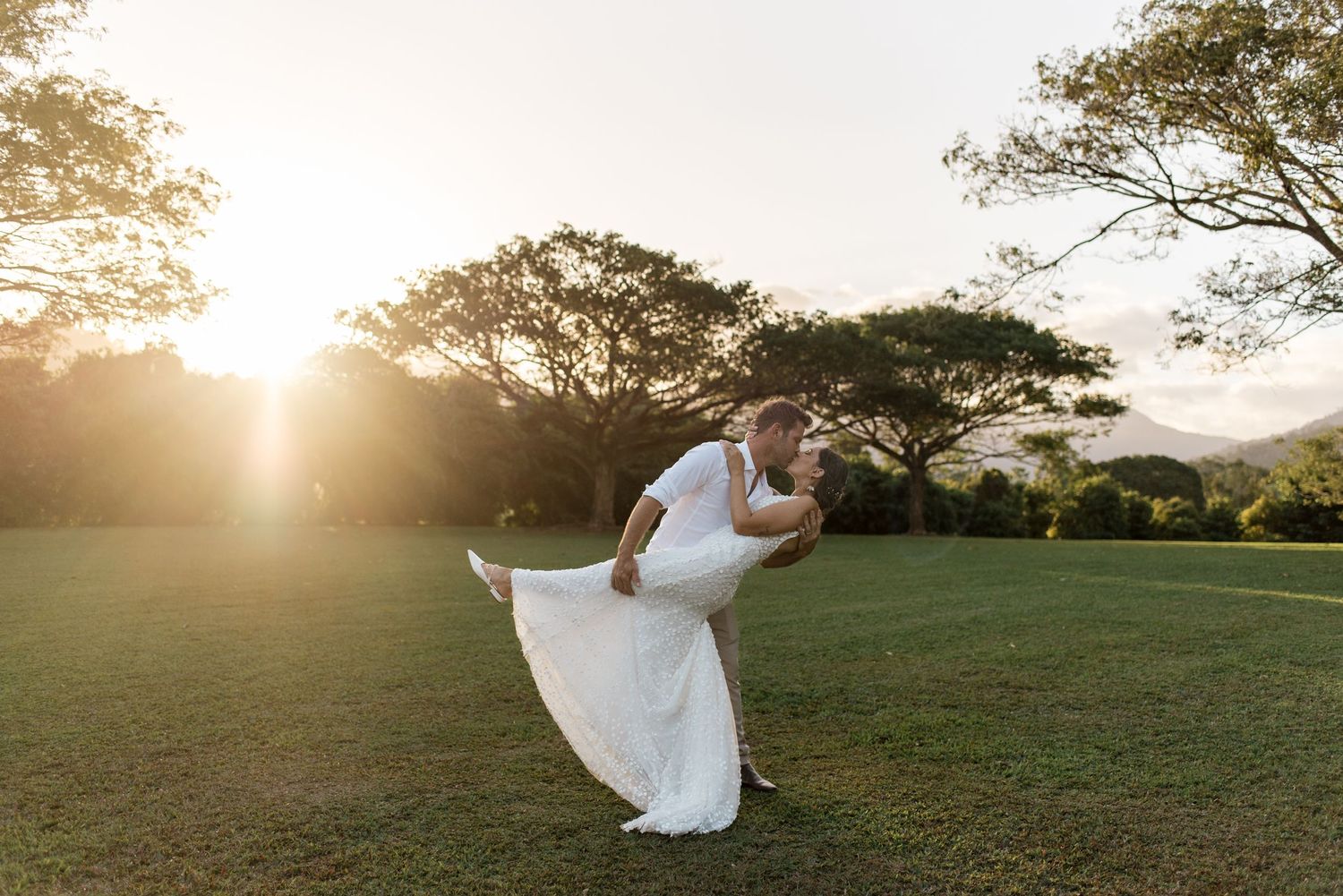 Groom dipping bride dramatically on green lawn with glowing sunset behind large trees.