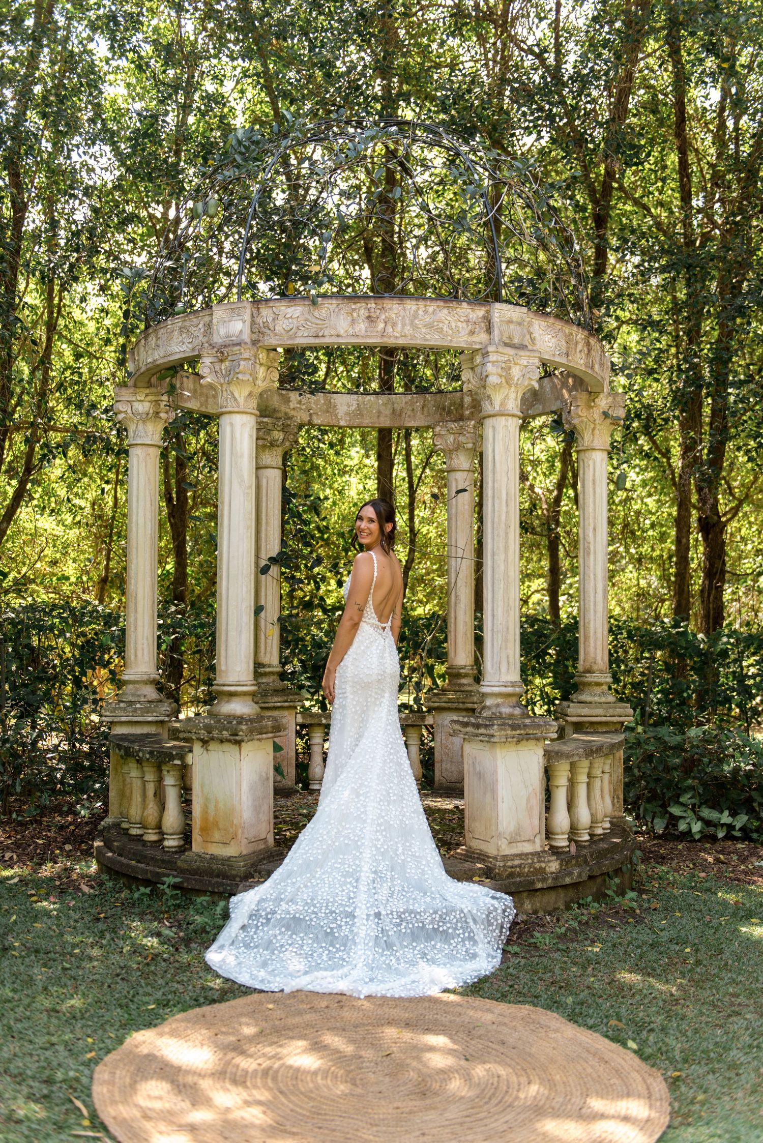 Bride in backless lace gown with cathedral train poses elegantly before ornate stone gazebo in garden.