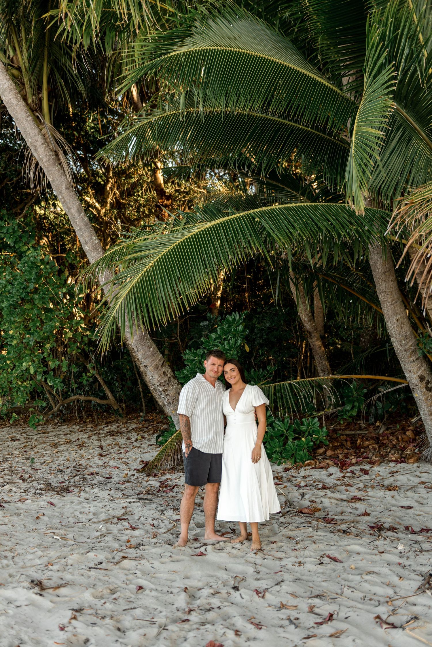 Couple standing barefoot on sandy beach surrounded by lush tropical palm trees during golden hour.