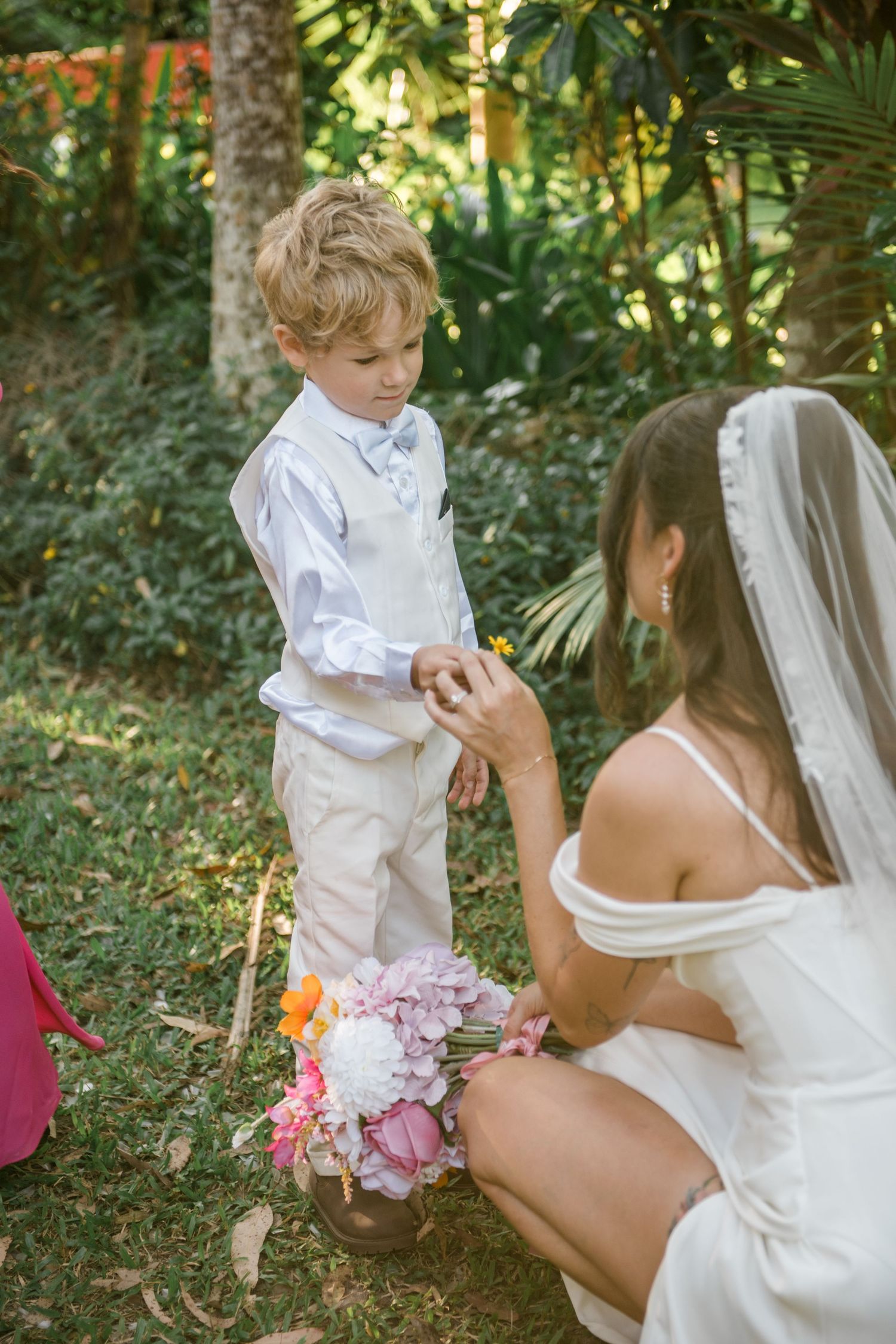Bride in white gown kneels to interact with young ring bearer in white suit outdoors.