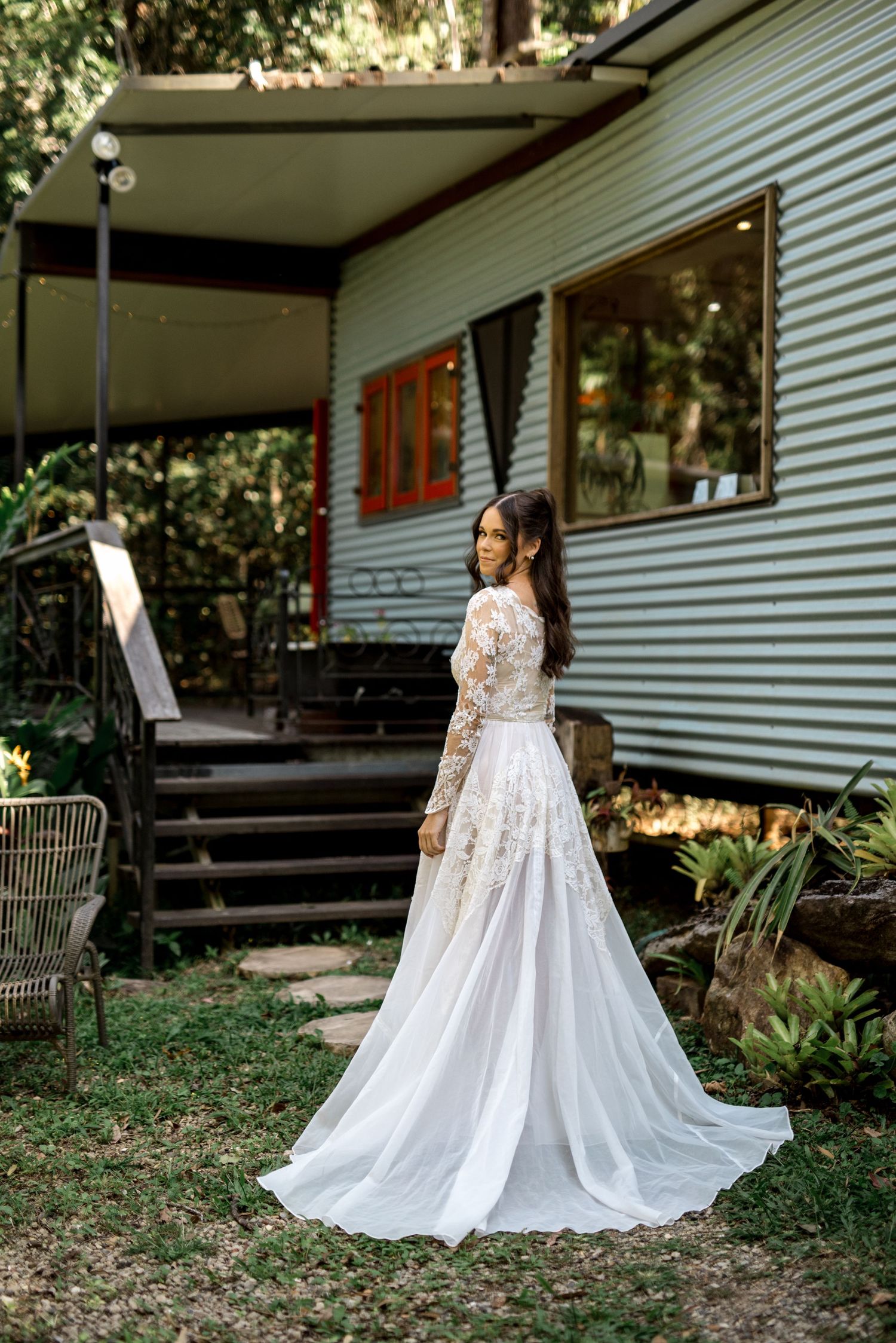 Bride in elegant long-sleeve lace wedding gown with flowing train standing outside a rustic tin cottage.