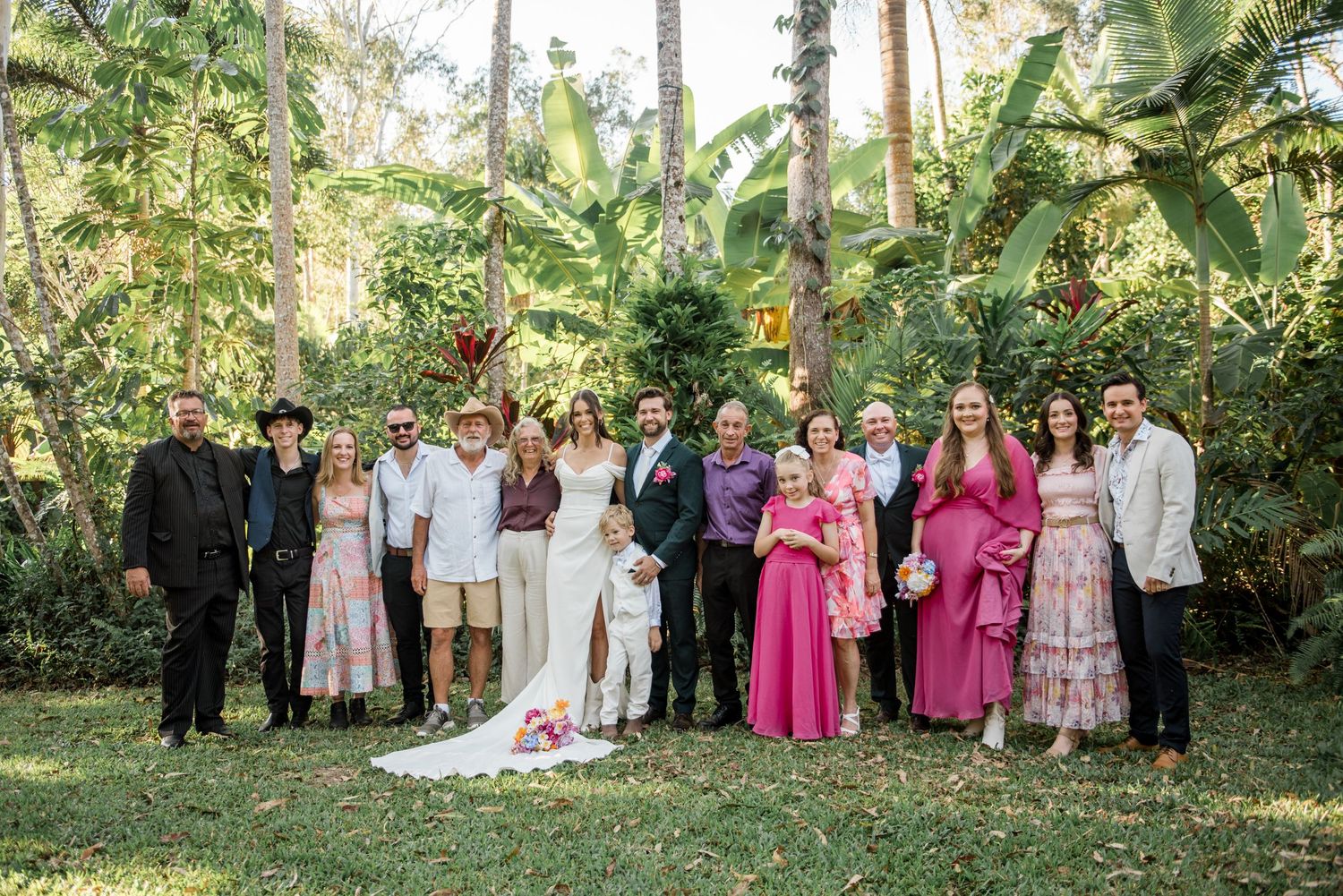 Wedding party group photo with bride and groom surrounded by family in a lush tropical garden setting.