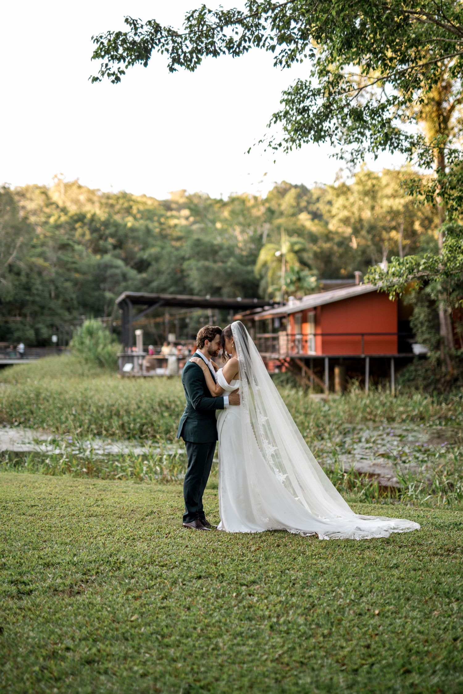 Bride and groom kiss tenderly on a green lawn with a red building and lush trees behind.