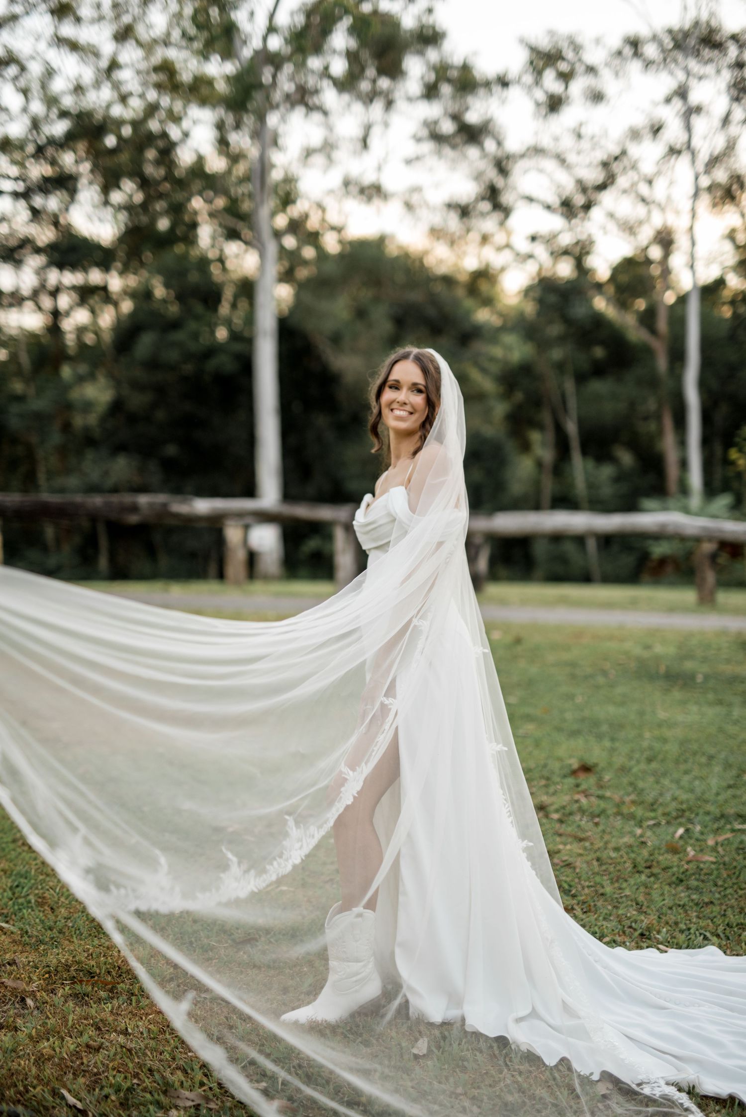 Bride in elegant white strapless gown with flowing lace-trimmed cathedral veil billowing in breeze outdoors.