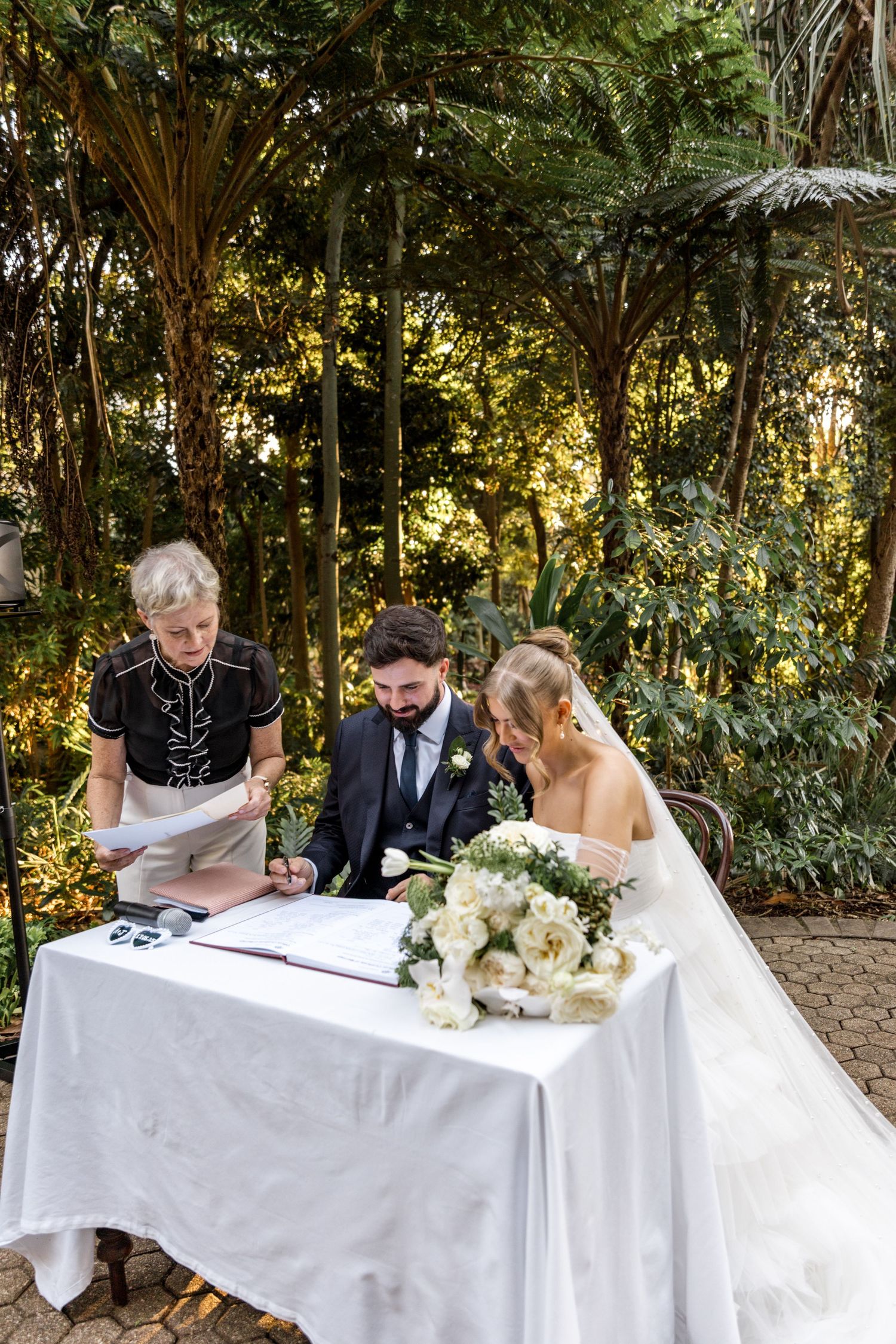 Bride and groom sign marriage certificate at outdoor garden ceremony with officiant and lush greenery.