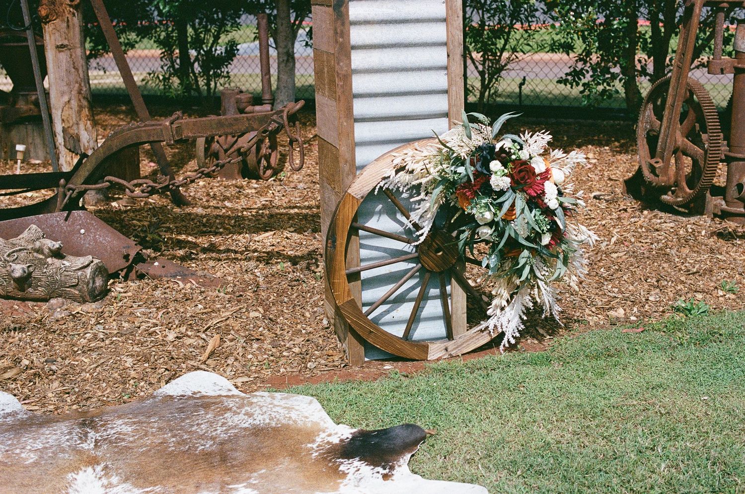 Rustic wedding decor featuring a wooden wagon wheel adorned with a floral wreath on mulched ground.