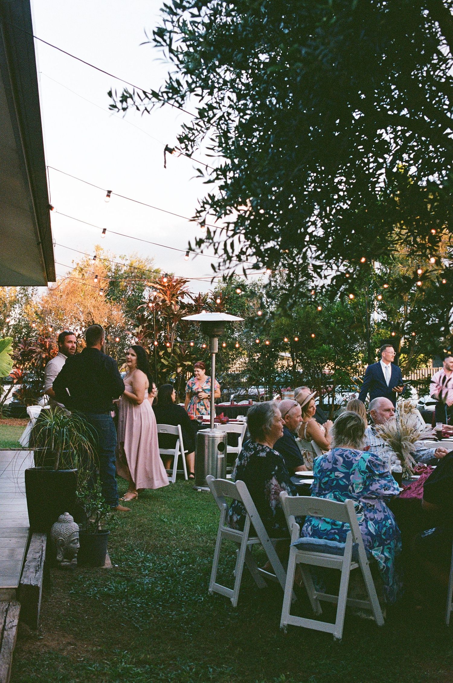 Wedding reception guests seated outdoors at dusk under string lights surrounded by lush garden greenery.