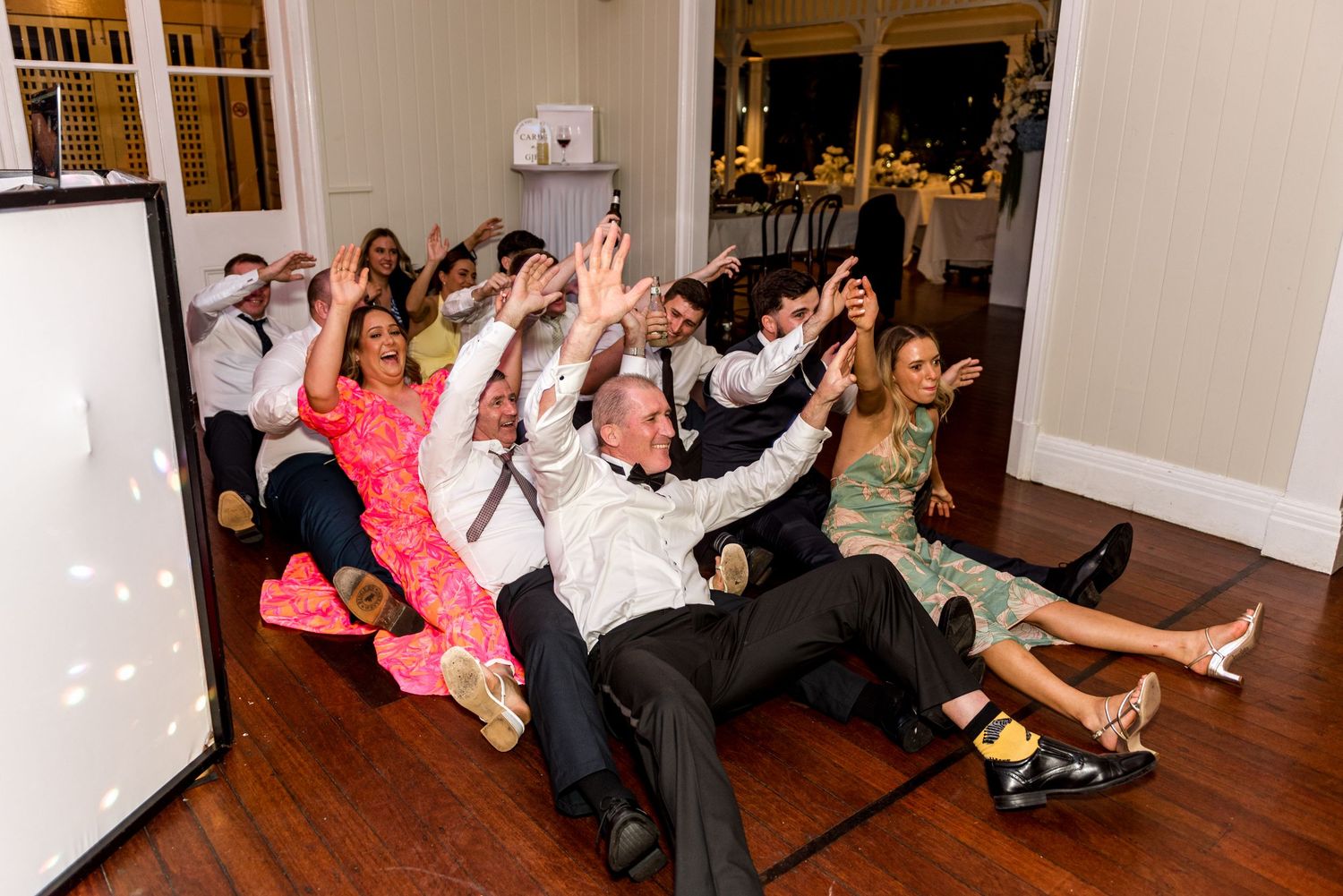 Wedding guests sitting on the floor with arms raised during a fun reception dance activity.