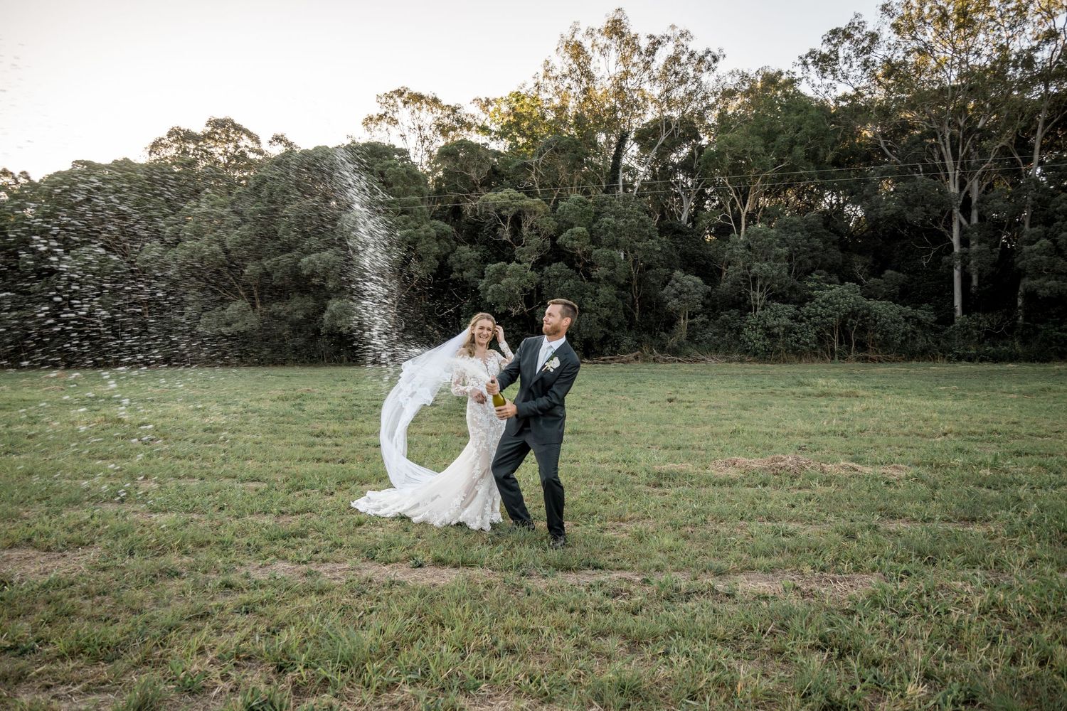 Bride twirls her long flowing veil in a sunlit green field as groom laughs beside her at sunset.
