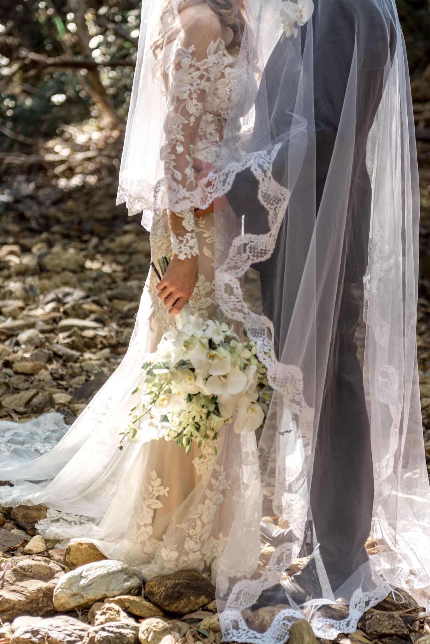 Close-up of bride in lace gown holding white bouquet standing beside groom on rocky ground.