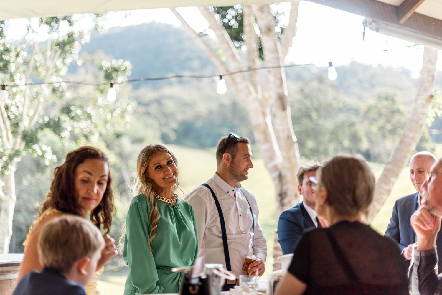 Wedding guests mingling under string lights on outdoor terrace with scenic green hills in background.