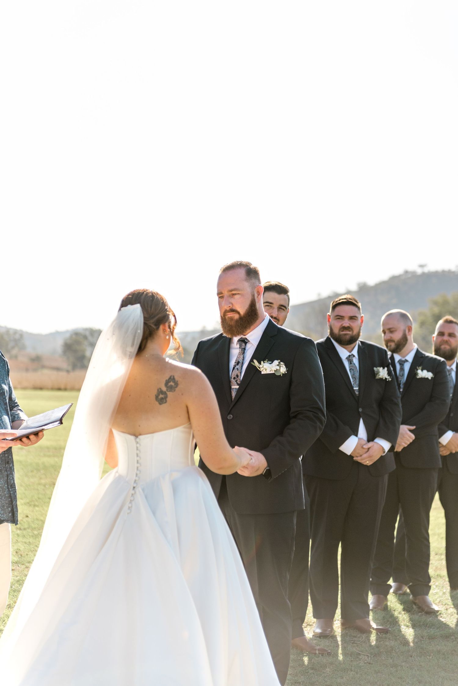 Groom with beard holds bride's hands at outdoor wedding ceremony with groomsmen standing in background.