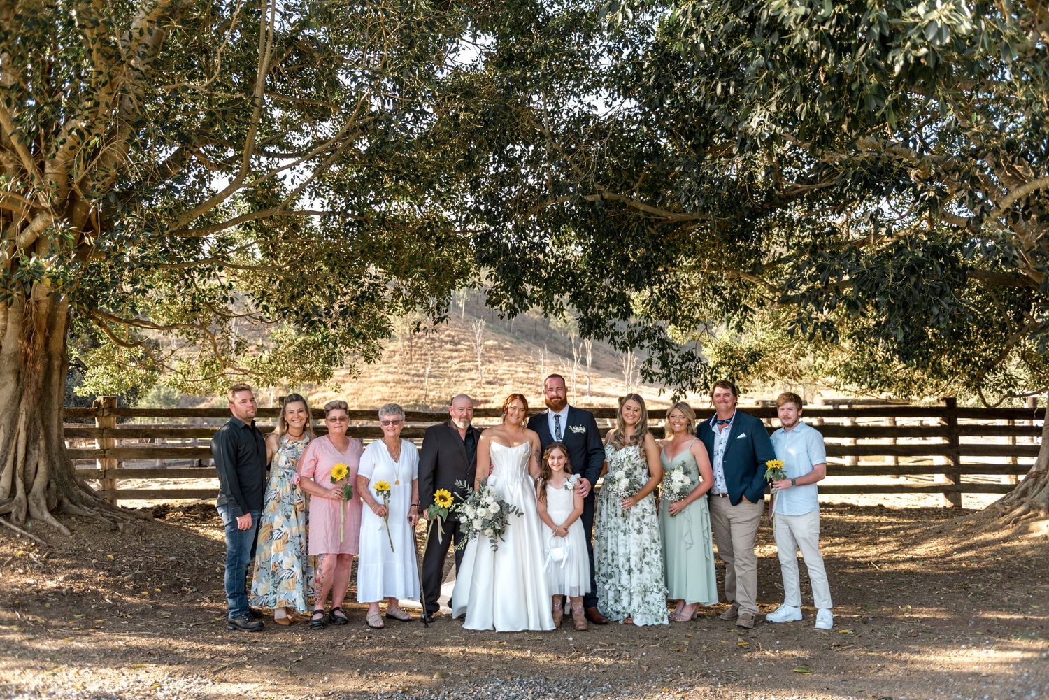 Wedding party group photo under large trees beside rustic wooden fence at outdoor venue.