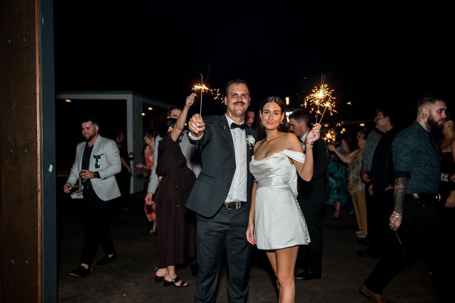 Newlywed couple poses with sparklers at outdoor evening wedding reception.
