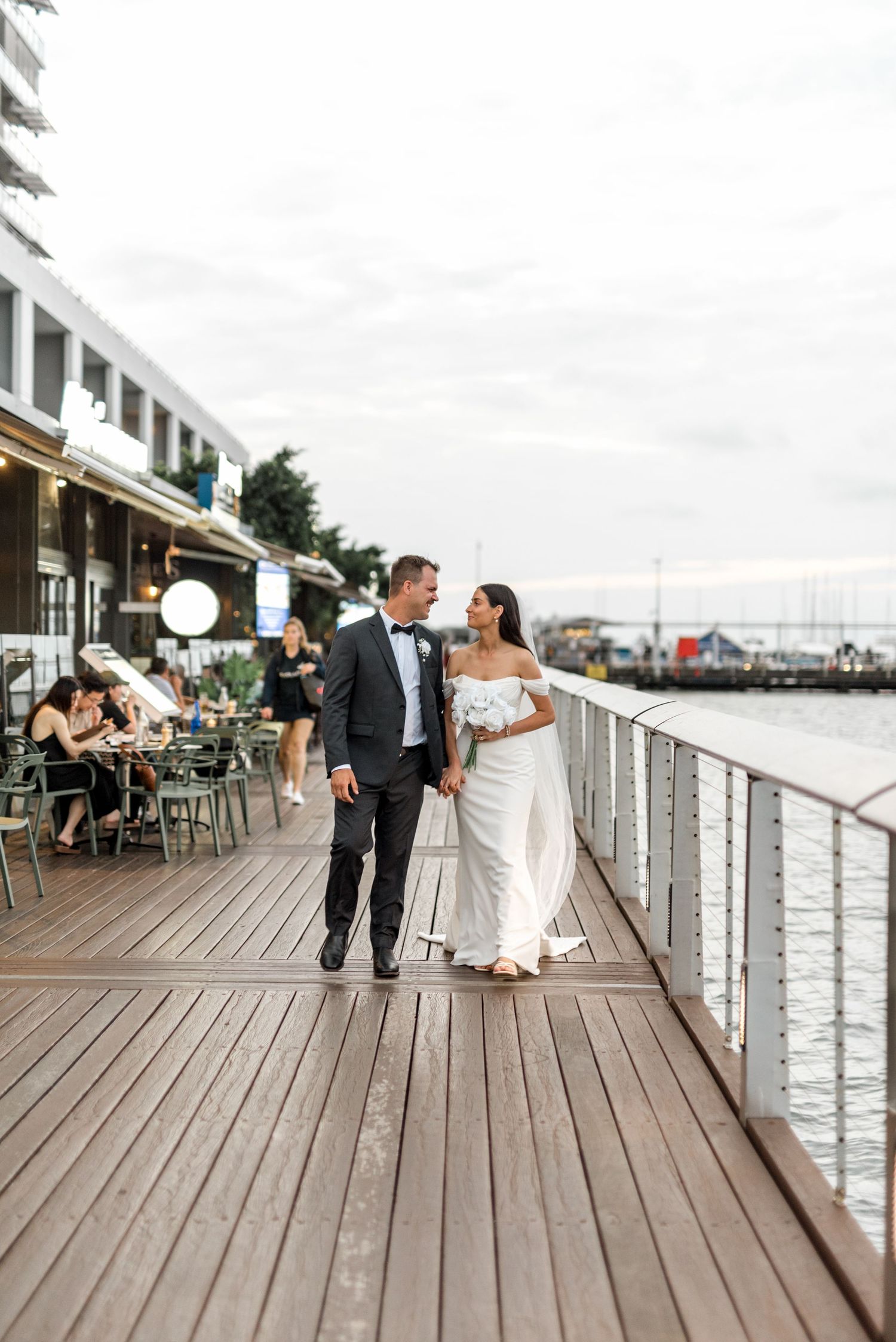 Newlyweds walk hand in hand along a scenic waterfront promenade on their wedding day.