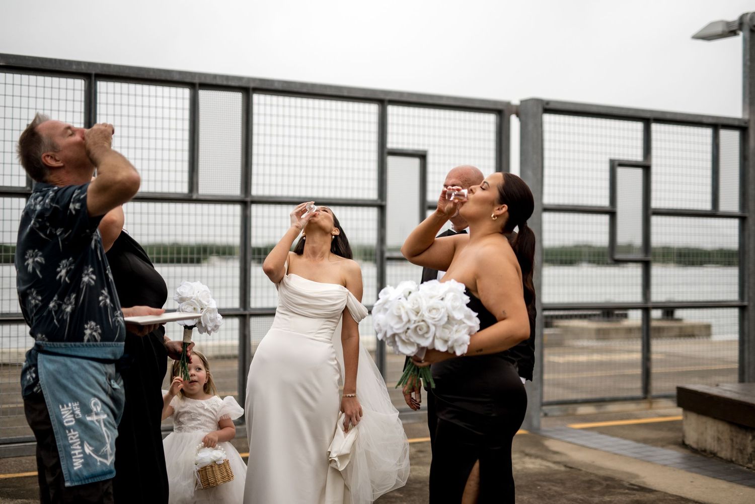 Wedding party doing shots outdoors near waterfront fence on a cloudy day.