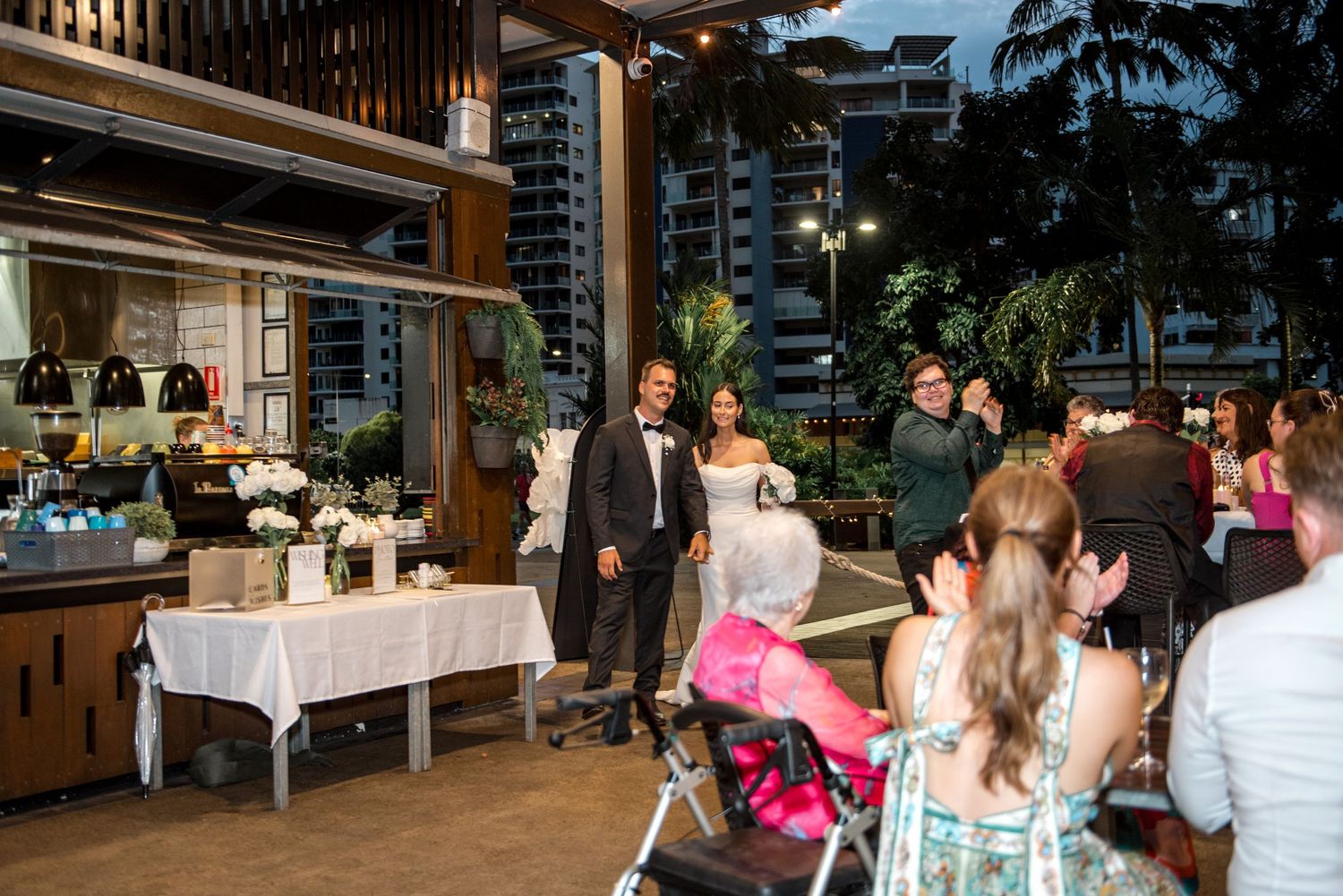 Bride and groom entering their outdoor waterfront wedding reception as guests applaud in the evening light.