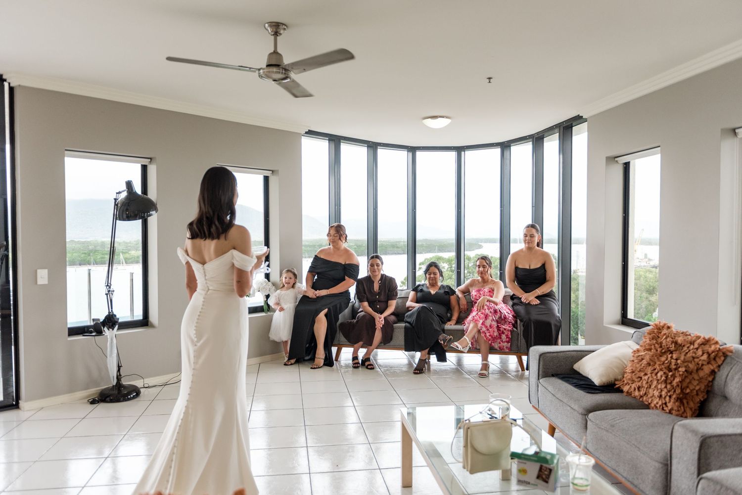 A bride in a white off-shoulder gown reveals her dress to emotional guests seated in a bright high-rise apartment.